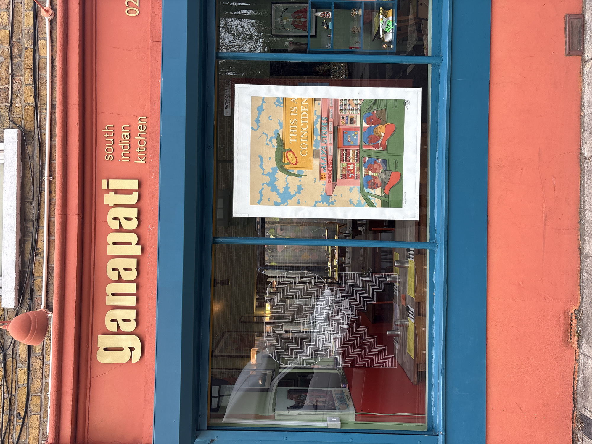The image presents the exterior of "Ganapati south indian kitchen," a restaurant located in London, United Kingdom. The building's upper facade consists of light-colored brickwork, beneath which is a coral-pink band. A terracotta-colored, bell-shaped light fixture protrudes from this band on the left. Gold, raised lettering spells "Ganapati," with smaller, dark letters below stating "south indian kitchen." The partial digits "02" are visible further to the right on this band.
Below, a teal-blue frame encloses the restaurant's large display windows. The left window pane reflects a white vehicle and exterior elements, while allowing visibility into the interior. Inside, dining tables are set with yellow placemats, cutlery, and metallic tumblers, against a red-painted wall. A prominent white decal, featuring an intricate, geometric, labyrinthine circular pattern, is affixed to the interior surface of this window.
The right window pane is largely obscured by a prominent vertical poster. This poster illustrates a green alligator-like creature, the partial text "THIS IS A COINCIDEN," a "MAYA STORES GROCERY" storefront, and a green car containing four stylized human figures. To the right of the poster, within the window's view, a teal-colored shelving unit displays various decorative objects, including small figurines and a framed item partially reading "MAKE YOUR OWN LUCK." The lower section of the building's facade, beneath the windows, is painted the same coral-pink and features a small rectangular vent above a grey paved sidewalk. No individuals are depicted directly in the scene.
