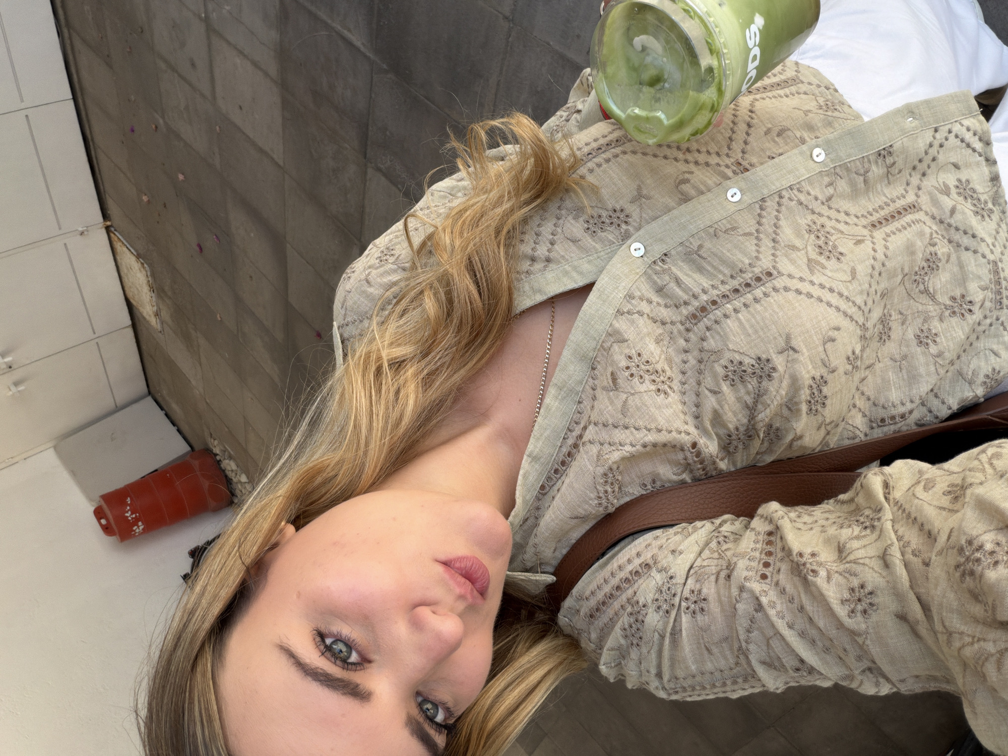 A selfie taken from an elevated angle shows a woman holding a green beverage. She has light skin, blue eyes, and blonde hair. She is wearing a light beige patterned shirt, and a brown leather strap is visible across her torso. The background includes a wall with a drain pipe and a section of flooring. The location is San Bernardino Tlaxcalancingo, Mexico.
