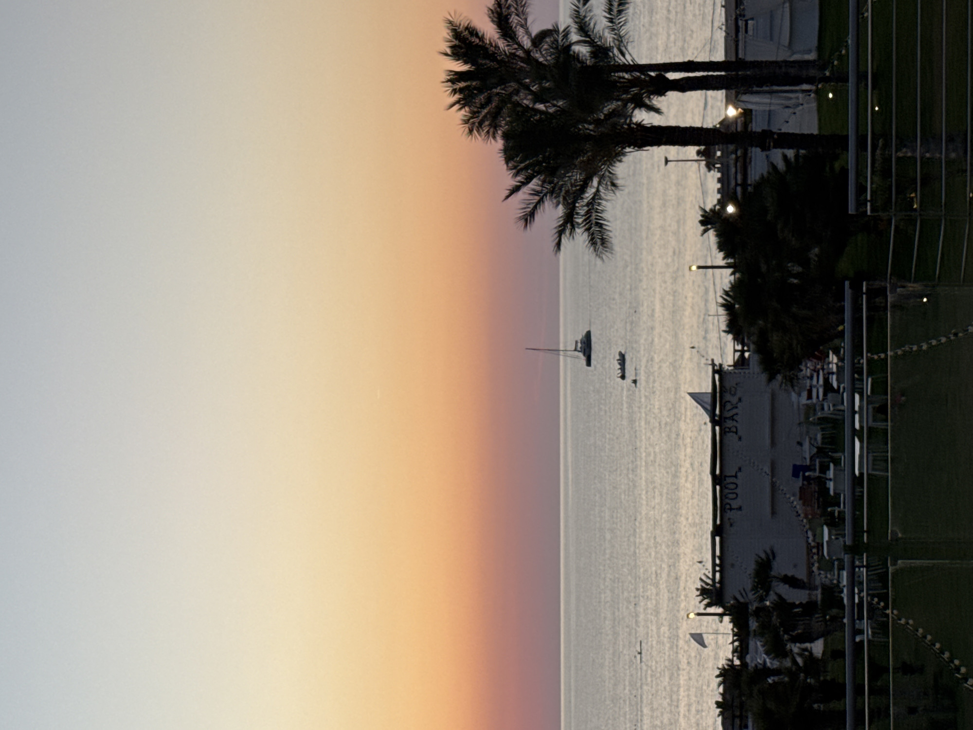 The image depicts a serene coastal scene at dusk in Isola delle Femmine, Italy. The sky transitions from a pale blue at the top to a soft orange and pink hue near the horizon, indicating the sunset. The calm sea stretches across the middle of the frame, reflecting the colors of the sky.
In the distance, several boats are visible on the water. A white sailboat with a single mast is positioned on the left side of the frame, while two smaller boats are closer to the center.
Along the shoreline, a structure with "POOL BAR" written on its facade is prominent. The building is white with a flat roof and appears to be part of a beachside establishment. Palm trees, silhouetted against the darkening sky, frame the right side of the image, their fronds casting shadows.
In the foreground, a dark grassy area is visible, separated by a metal railing. The overall atmosphere is peaceful and tranquil, suggesting a quiet evening at the seaside.