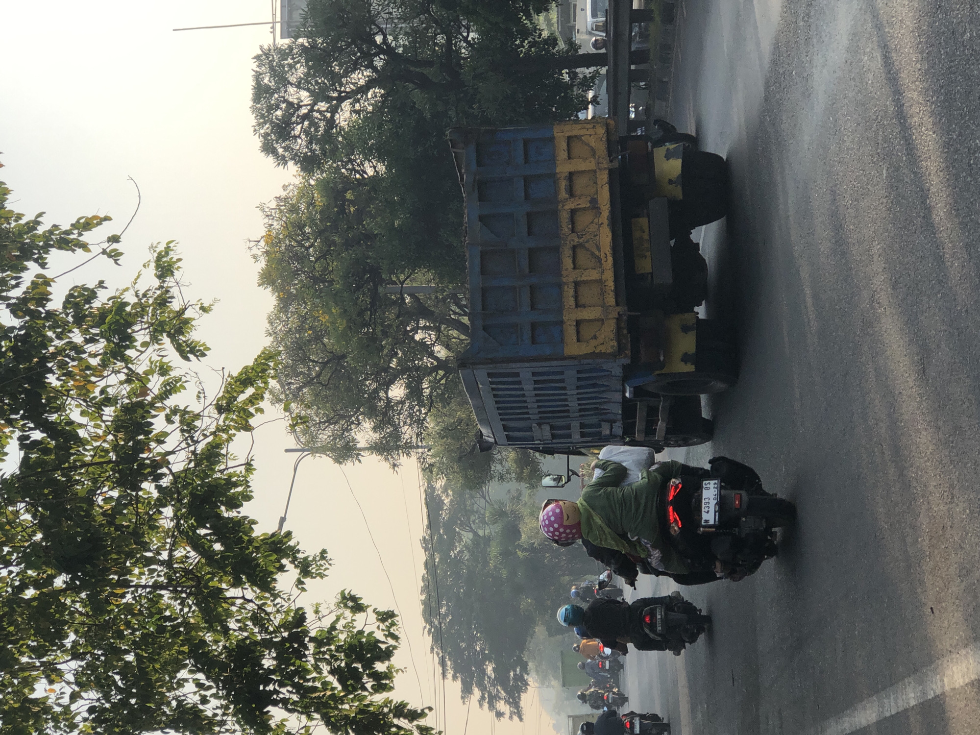 In Kludan, Indonesia, on a hazy morning, a truck and several motorcycles navigate a two-lane road. The truck, a blue and yellow dump truck, is in the right lane, facing away from the camera. The motorcycles are in the left lane, also facing away. One motorcycle in the foreground carries two individuals. The person in the back, wearing a green jacket and a pink helmet with a face pattern, appears to be a child. The road is asphalt, marked with a white line on the left side. Trees with green foliage line the sides of the road, and streetlights are visible in the background. The sky is bright and overcast.