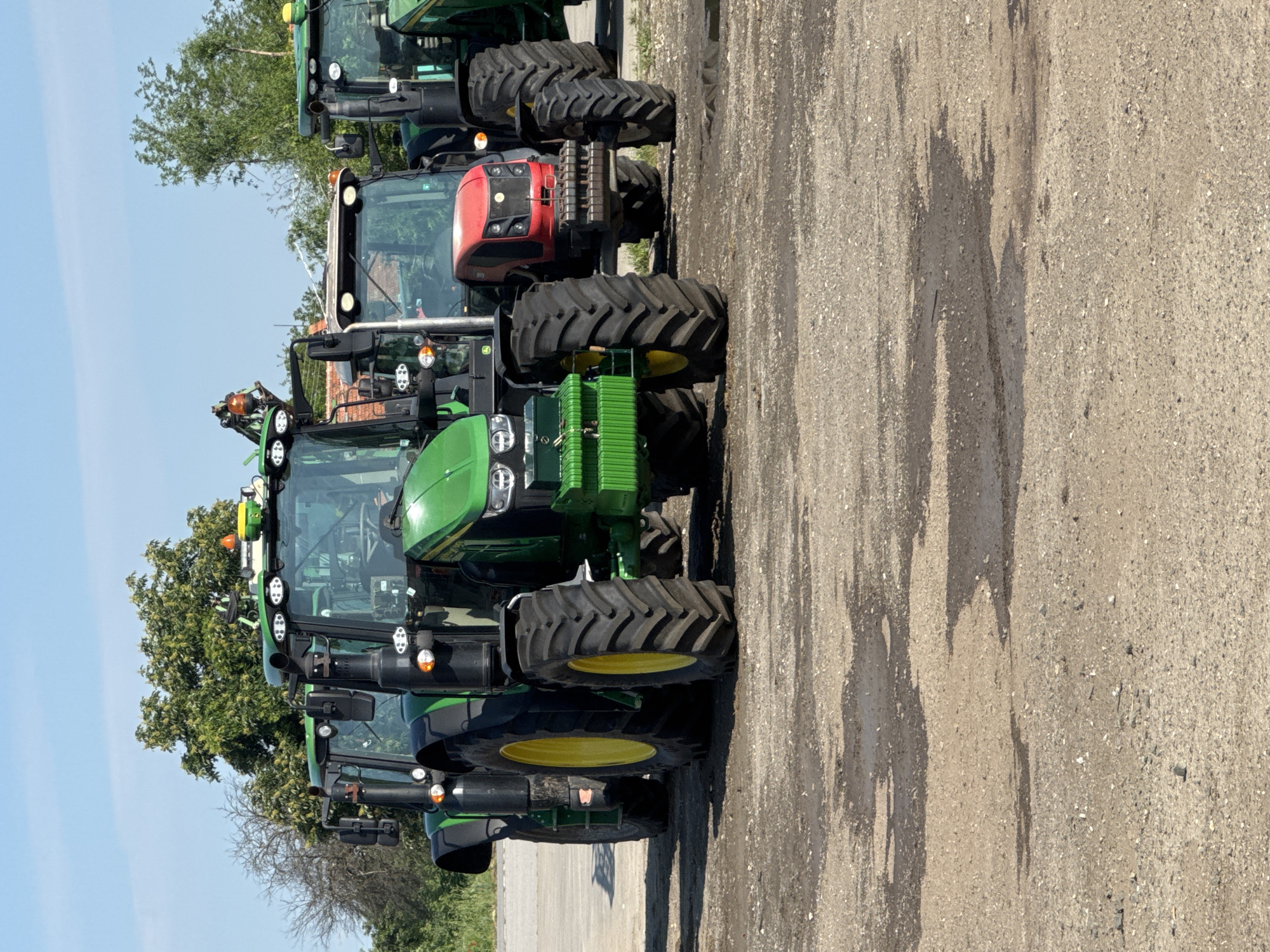 The image shows a row of parked tractors on a concrete surface. The tractors are primarily green, with one visible red tractor. The location is unknown, and there is no information available to verify its location in Sarnevo, Bulgaria. The environment is clear with a blue sky and visible trees in the background. There are no people visible in the image.