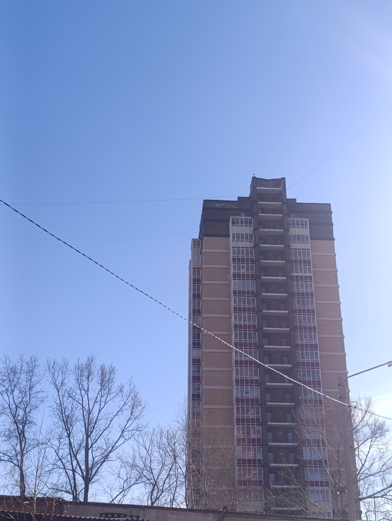 The image captures a high-rise apartment building in Krasnoyarsk, Russia, on a clear day with a blue sky. The building has a facade with balconies, and its construction includes brick and glass. Power lines stretch across the sky in front of the building. Below the building, bare trees and a rooftop are visible. There are no people visible.