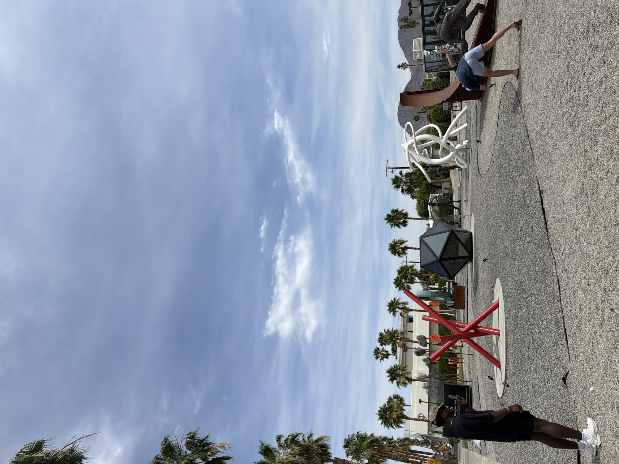 The image captures a scene in Palm Desert, California, United States, likely along El Paseo Drive, known for its public art displays. The photograph is taken from ground level, looking towards a row of palm trees flanking the street, against a backdrop of a clear, blue sky with scattered clouds.
Several sculptures are visible along the walkway. A red abstract sculpture comprised of beams rising from a circular base, as well as a black geodesic dome and a complex, white swirling sculpture. Towards the background of the image, a man is seen in a handstand. A dark-skinned man in a black shirt, shorts, and sneakers, wearing a hat and holding a mobile phone, appears to be taking a photograph. The walkway is covered in light-colored gravel.