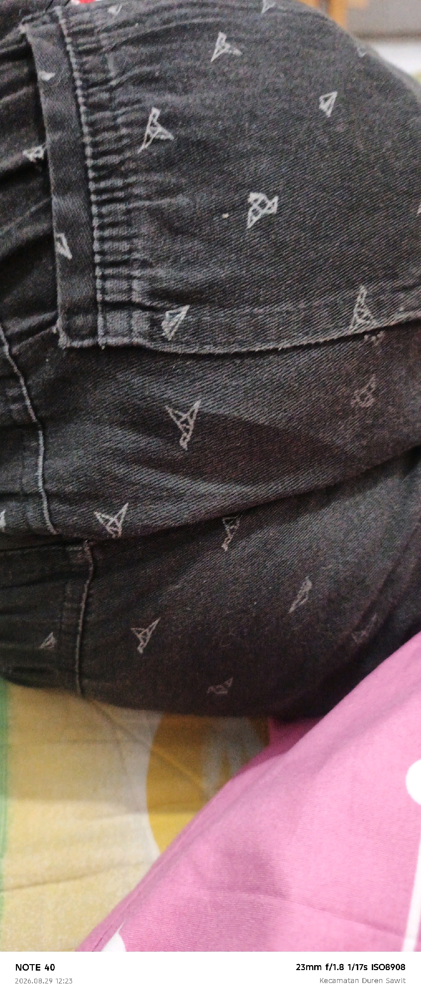 The image shows a close-up view of dark denim fabric with a subtle repeating geometric pattern of stylized triangles. The fabric appears to be part of a garment, possibly pants, with visible stitching and seams. The texture of the denim is detailed, showing the weave of the material. In the lower portion of the image, the fabric transitions to a pink textile, which is slightly out of focus. The bottom edge of the image contains text overlay, which reads "NOTE 40 2026.08.29 12:23" and "23mm f/1.8 1/17s ISO8908 Kecamatan Duren Sawit". The presence of "Kecamatan Duren Sawit" indicates the location as Duren Sawit District, East Jakarta, Indonesia.