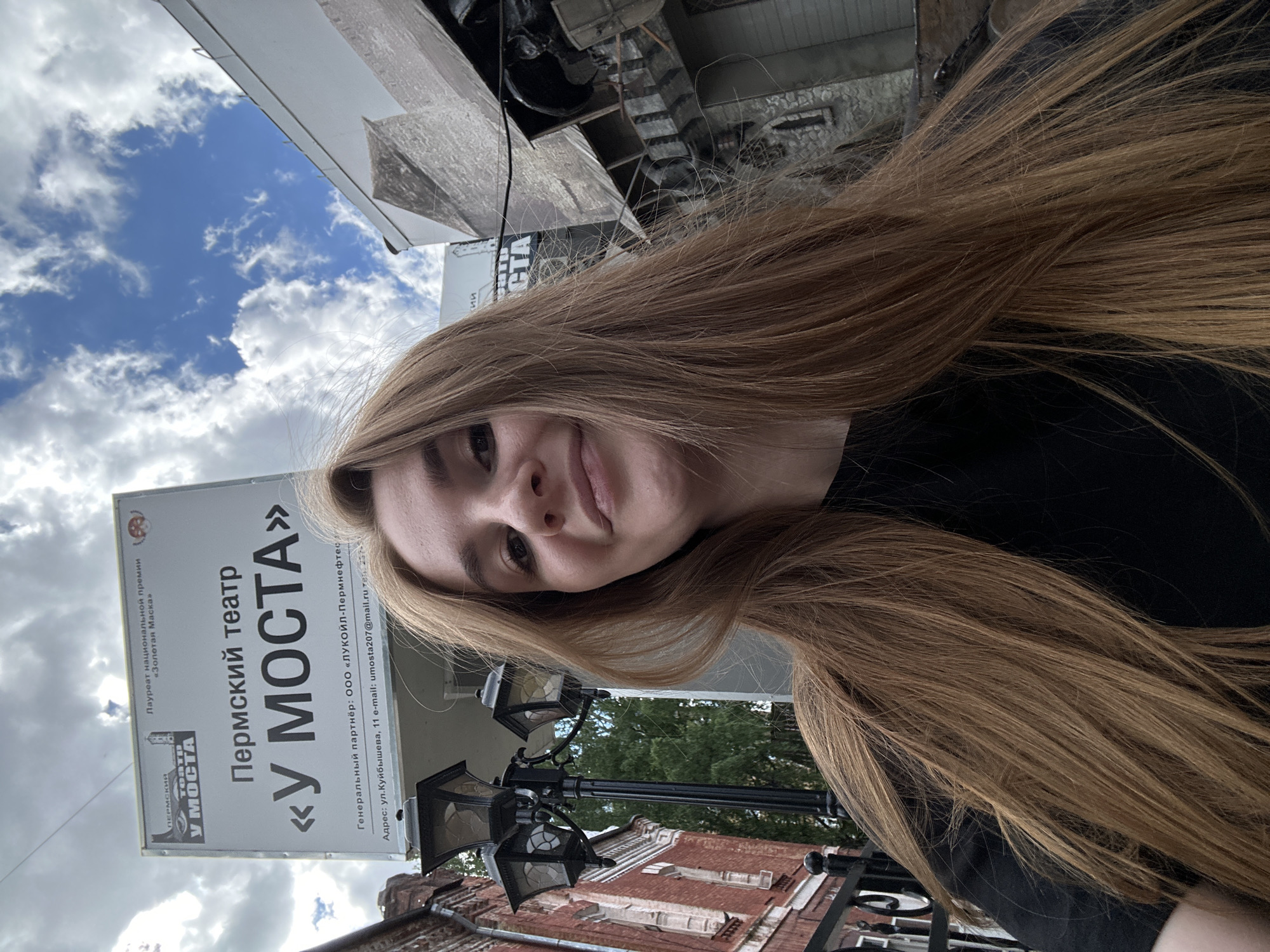 A young woman with long, reddish-blonde hair smiles at the camera, taking a selfie. She is wearing a black t-shirt. In the background, a sign for the "Permian Theater 'U Mosta'" is visible, along with a brick building and a street lamp. The sky is blue with scattered white clouds. The image appears to have been taken on a sunny day in the city of Perm, Russia.