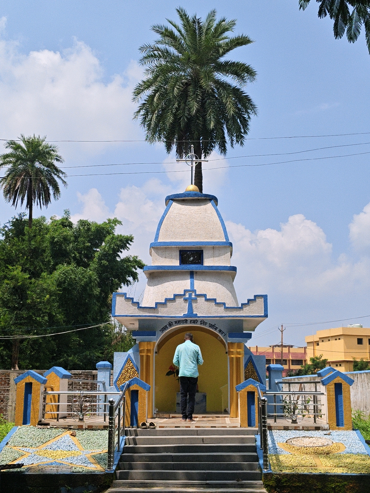 In Mandar, India, a person in a light blue shirt and dark pants stands on the steps of a small, ornate structure adorned with blue and yellow accents. The structure has a conical roof and a large palm tree grows directly out of its apex. The scene is set against a backdrop of a bright blue sky with scattered white clouds. To the left of the structure, lush green trees are visible, and further in the background, several buildings can be seen, one of which is yellow. The ground in front of the structure is decorated with colorful mosaic patterns, including a prominent star design. Two pairs of sandals are placed near the entrance.
