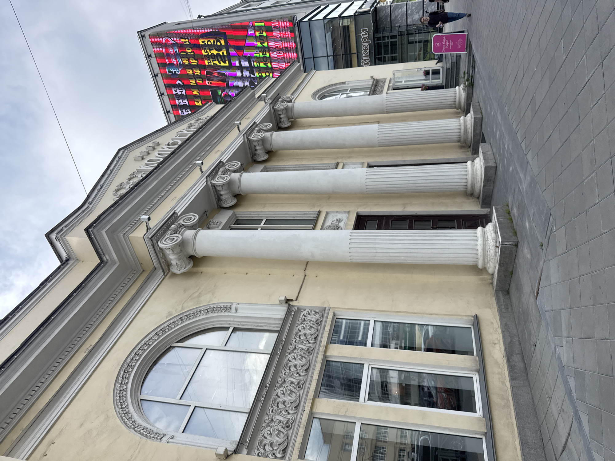 The image shows the exterior of a building with classical architectural elements in Yekaterinburg, Russia. The building has a pale yellow facade with large arched windows and a series of white ionic columns. Above the columns, there is ornate decorative molding. To the right of this building, a modern glass-fronted building is visible, topped with a large, brightly lit digital billboard displaying colorful, abstract patterns and text in Russian. In the foreground, a paved sidewalk with a patterned tile design runs in front of the building. A few people are visible on the sidewalk; one person, wearing dark clothing, is walking away from the camera, while another individual in a dark t-shirt and jeans is walking towards the right edge of the frame. A pink directional sign stands on the sidewalk near the entrance of the building. The sky is overcast. The digital billboard is advertising a mobile phone company, with text visible including "Аптека" (Apteka - Pharmacy) and "ЕМИШОП" (EMISHOP). The building with the columns appears to be the "Dom Kino" (House of Cinema) located at 139 Lenina Street, Yekaterinburg.