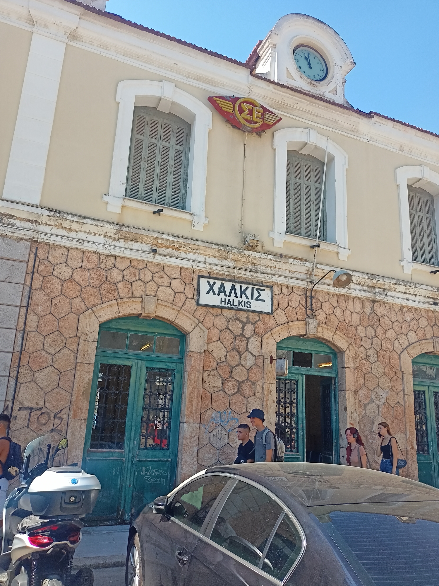The image captures the exterior of a train station in Chalkis, Municipal Unit of Anthidon, Greece. The station building features a cream-colored upper facade with arched windows and white trim, topped by a red-tiled roof and a decorative clock. Below the windows, the building is constructed with a distinctive hexagonal stone pattern. A sign prominently displays "ΧΑΛΚΙΣ" and "HALKIS" in black lettering on a white background. Two teal-colored doors with ornate metal grilles are visible, leading into the station.
In front of the station, several people are present. Two young men and a woman with headphones are standing near an open doorway. Another woman, wearing a black tank top and jeans, is talking on her phone. To the left, a motorcycle with a white top box is parked on the sidewalk. A dark-colored car is partially visible in the foreground, obscuring some of the lower portion of the station. Graffiti is present on the stone facade of the building. The sky is clear and blue, indicating a sunny day.