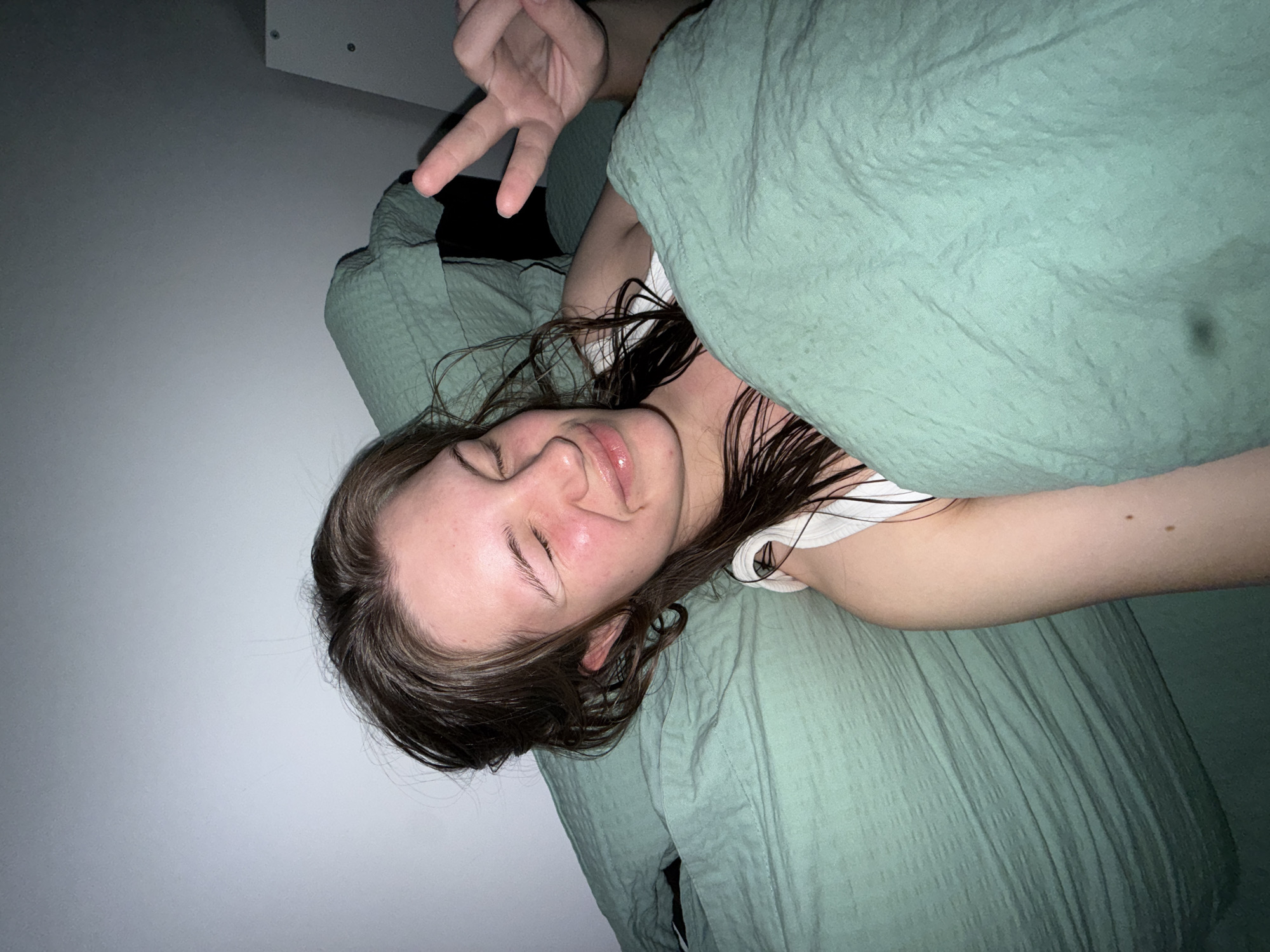 A person is lying on a bed covered with a green duvet. Their head is tilted towards the viewer's left. They have brown hair, closed eyes, and appear to be smiling slightly. Their right hand is visible in the upper left corner of the image, making a peace sign. They are wearing a white top. The background consists of a light gray wall. The location is not evident in the provided image.