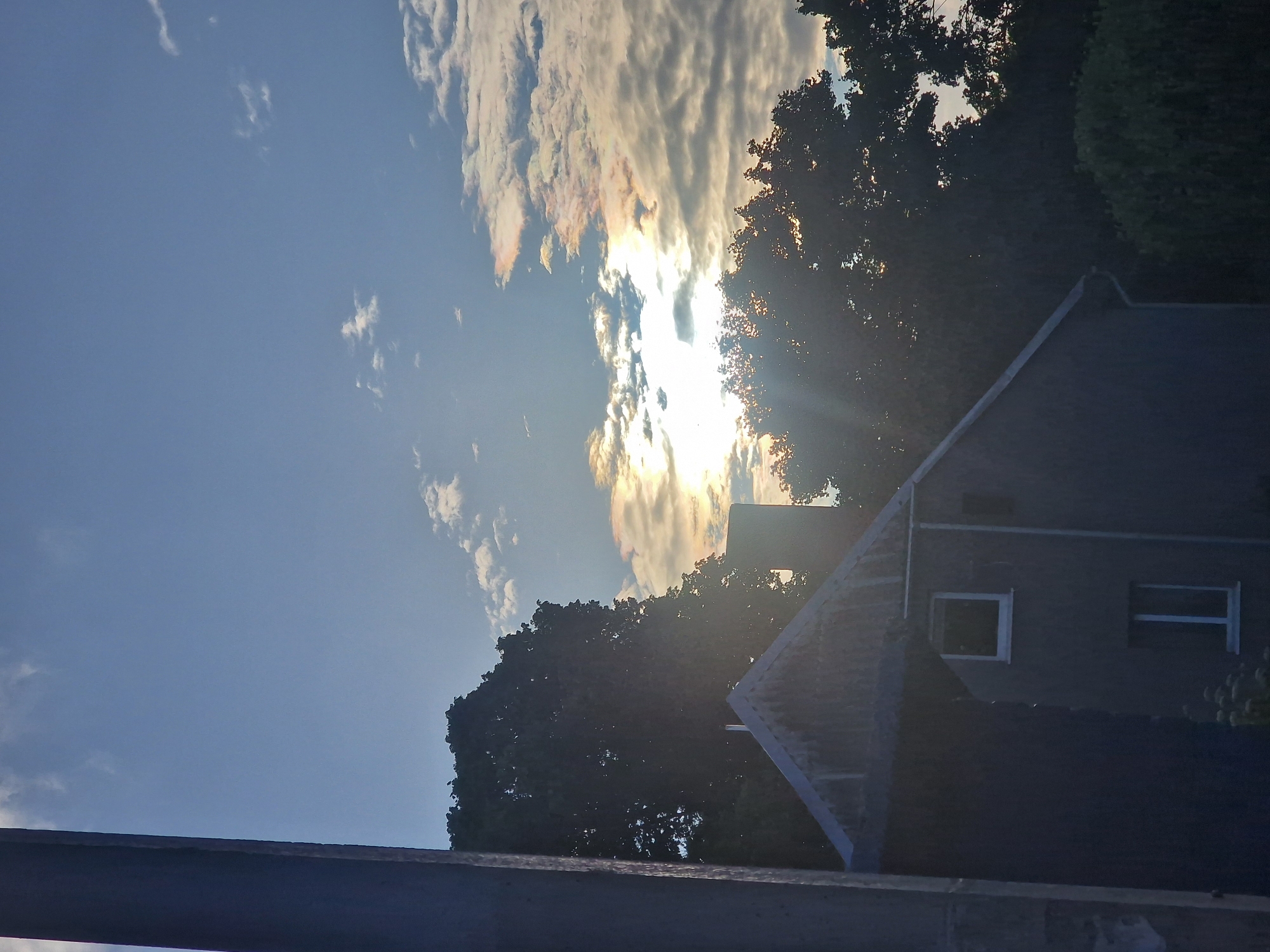 An exterior, eye-level, vertical, medium shot captures a silhouetted house with a pitched roof in the foreground, partially obscured by a dark, vertical structure on the left. The house has two visible windows with white frames, one square and one rectangular. Behind the house, silhouetted trees create a dense backdrop against a sky with scattered clouds. The sun is visible through the clouds, casting a bright, lens-flare effect and highlighting iridescent colors within the cloud formations. The overall scene is dominated by a gradient from deep blue in the upper sky to lighter hues near the horizon, with a twilight or sunrise/sunset ambiance. The location is specified as Романово, Russia.