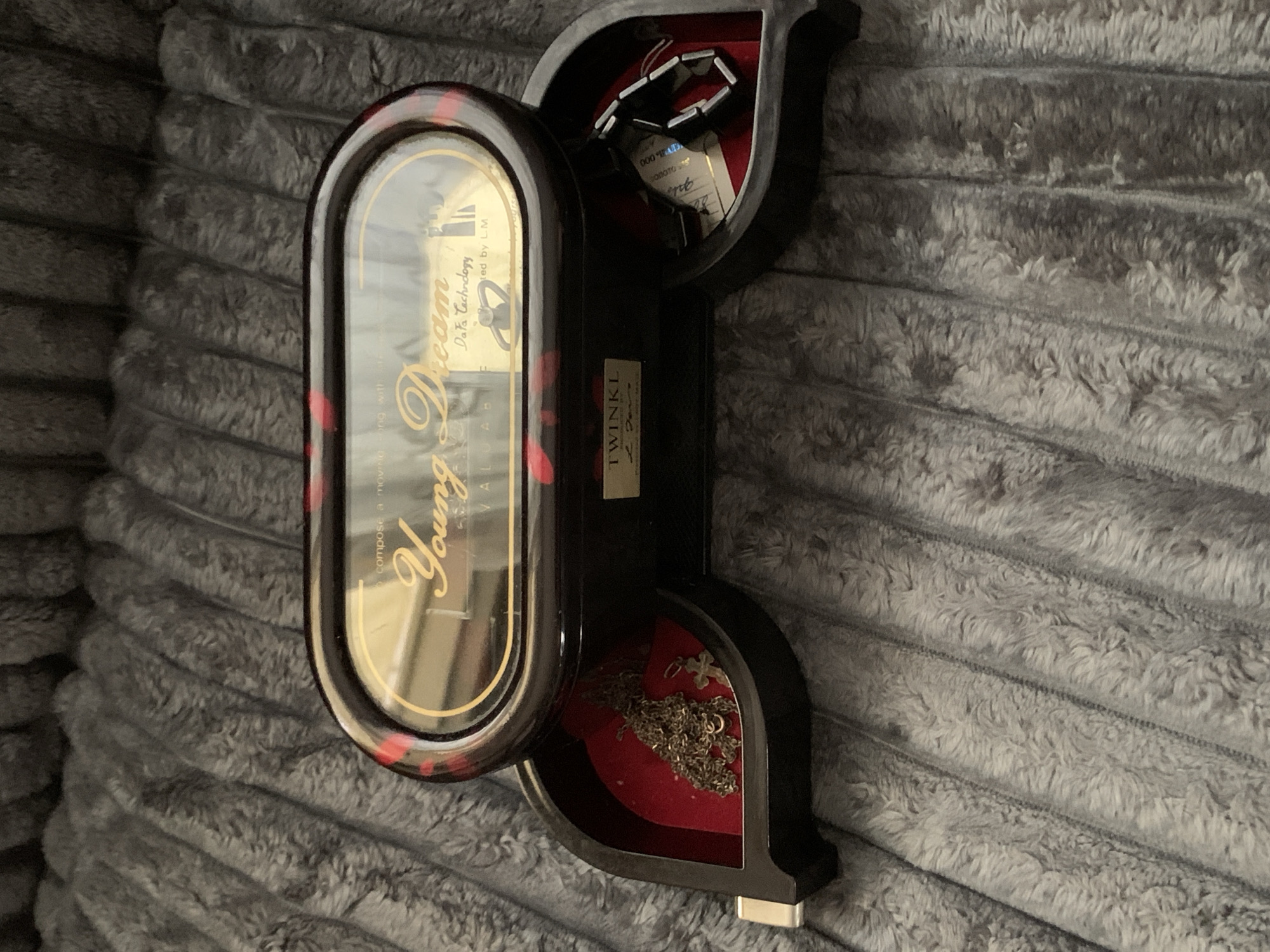 A dark, lacquered jewelry box with a red and black marbled lid sits open on a gray, plush blanket. The lid is oval and transparent, revealing a gold-toned interior with text that reads "compose a moving song with a dream" and "Young Dream." Below this, smaller text indicates "VALIABLE" and "Data Technology," with what appears to be a music box mechanism. The main body of the box is black with red accents. The jewelry box has two open drawers. The left drawer contains a silver-colored chain necklace with a cross pendant. The right drawer holds a black bracelet. The blanket appears to be the primary surface the box is resting on. No city or location information is discernible from the image.