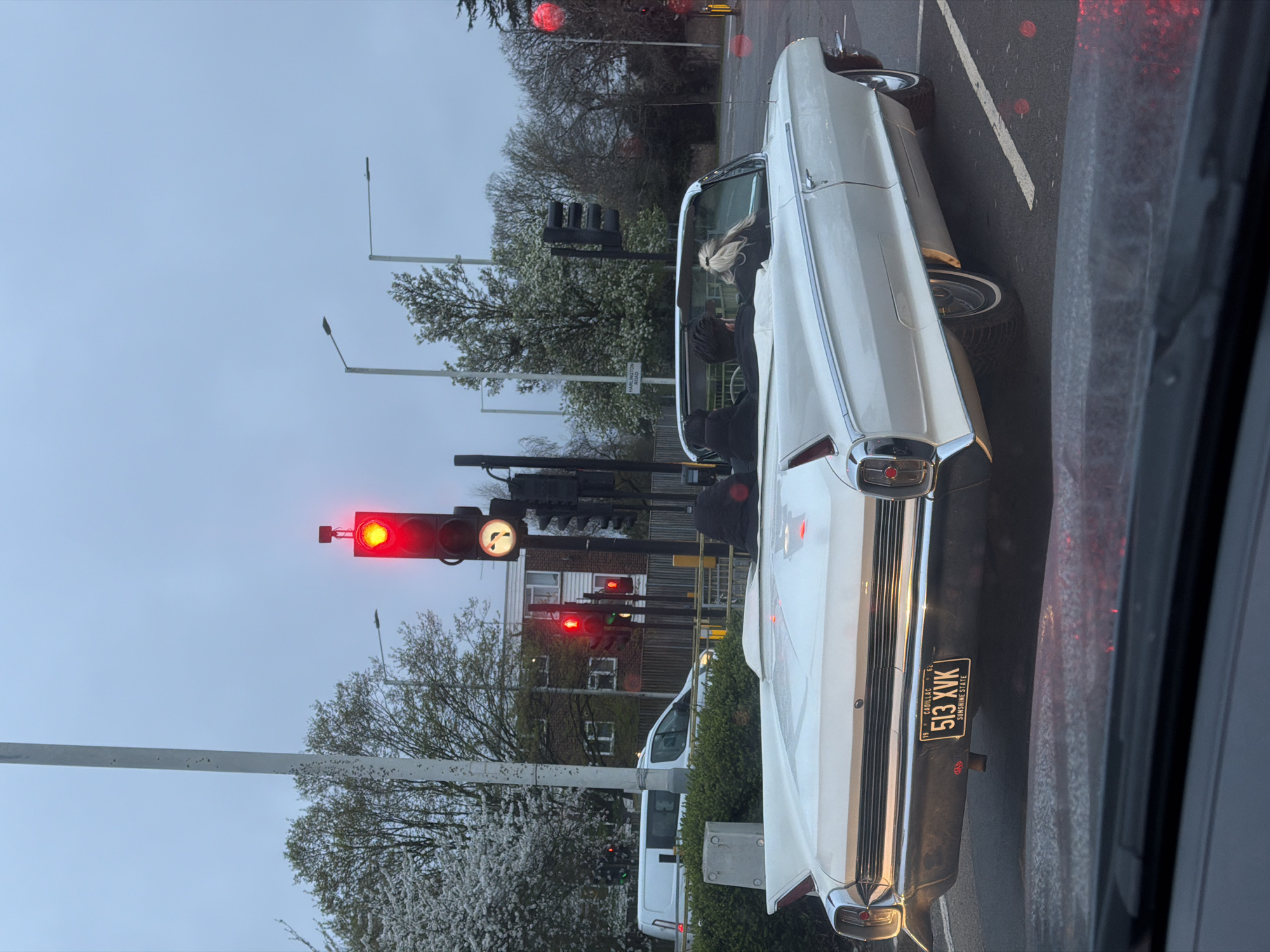 The image shows a white 1962 Cadillac convertible stopped at a traffic light. The license plate reads "513 KVK," identifying it as registered in the "Sunshine State." The traffic lights are red. The image was taken in Harlington, London, United Kingdom. There are at least two people in the car, one of whom has blond hair. The street has flowering trees and street lights. A white van is visible behind the car. The image was taken from inside another vehicle.
