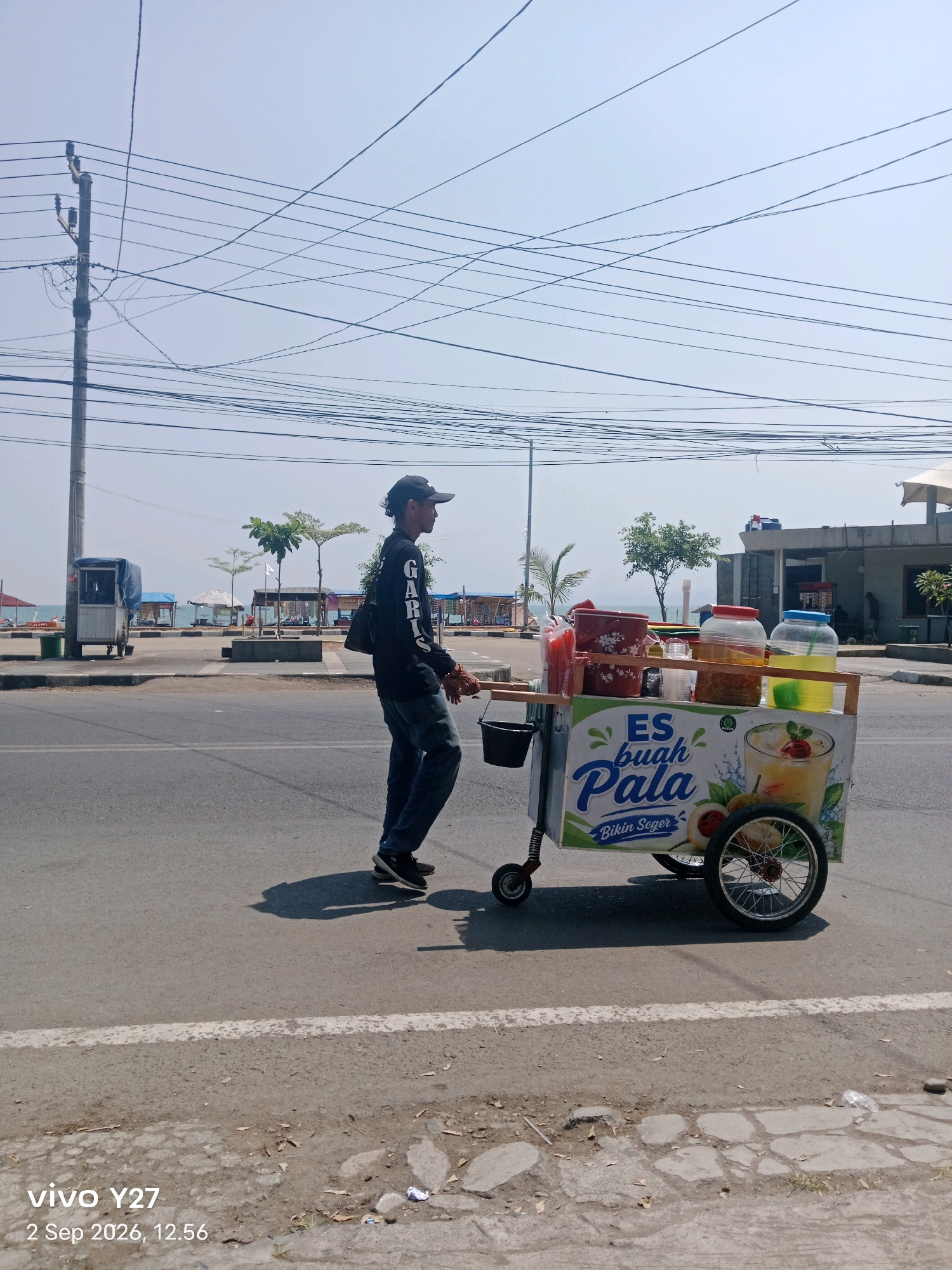 A man walks on the right side of a paved road in Palabuhanratu, Indonesia, pushing a cart filled with drinks. The man is wearing a black long-sleeved shirt with "GARIS" printed on the left sleeve, blue jeans, and a black baseball cap. He is carrying a black bag over his right shoulder and holding the cart's handle with his right hand. The cart is decorated with images of fruit and text that says "ES BUAH PALA Bikin Seger". It contains various containers of colorful beverages, ice, and what appear to be fruits. In the background, there is a utility pole with many overhead wires, some trees, a beach with umbrellas, and buildings. The sky is clear and blue. A watermark in the lower left corner of the image reads "vivo Y27 2 Sep 2026, 12.56".