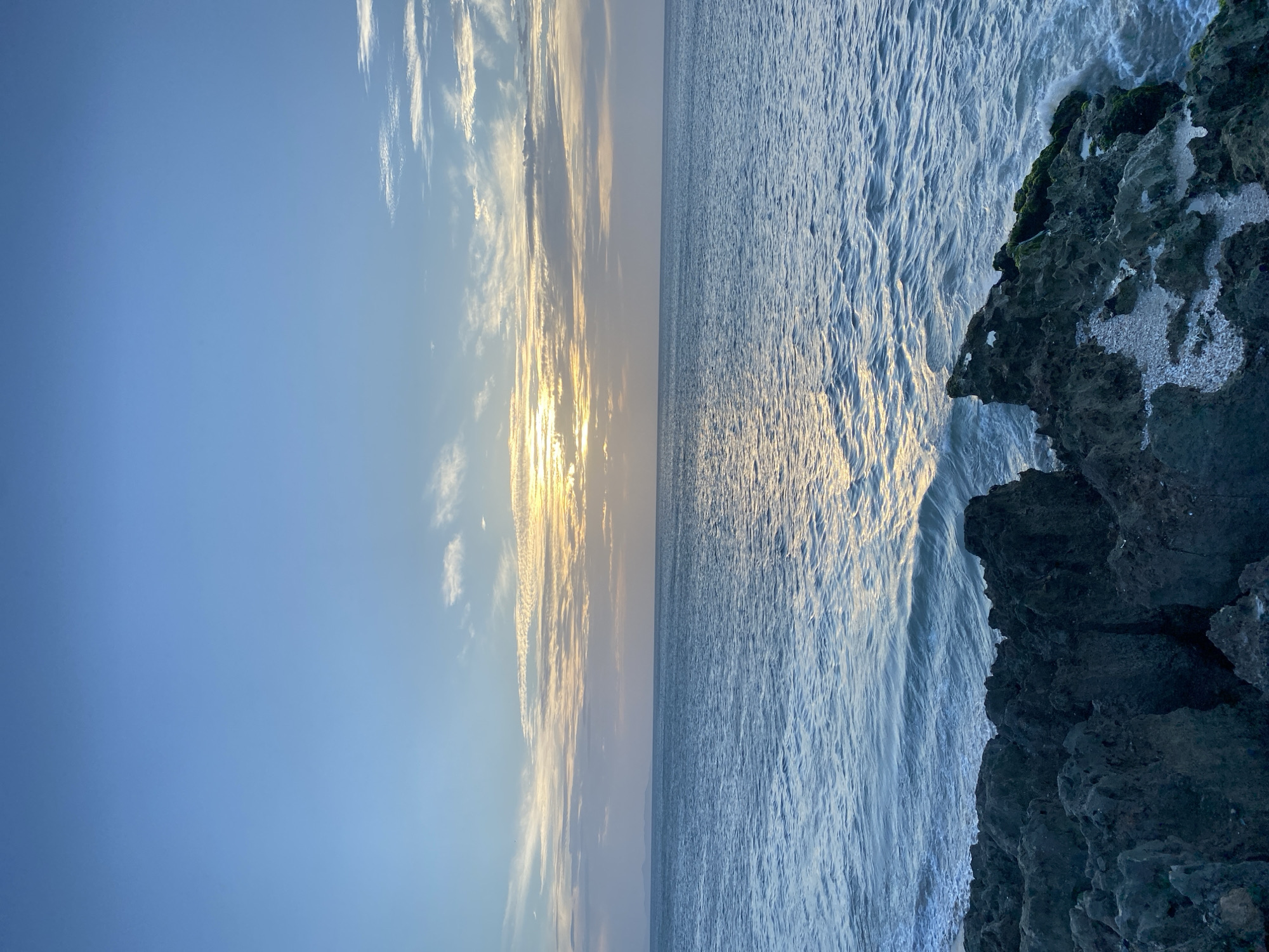 The image shows a seascape in Juan Griego, Venezuela, likely taken from a rocky coastal area. The sky, partially obscured by clouds, transitions from a deep blue to a lighter hue as it nears the horizon where the sun is setting. The water reflects the sunlight, creating a shimmering effect. On the right side of the image, there is a dark, craggy rock formation, part of the coastline, which is partially covered with what looks like wet areas from splashing waves. The scene is devoid of people.