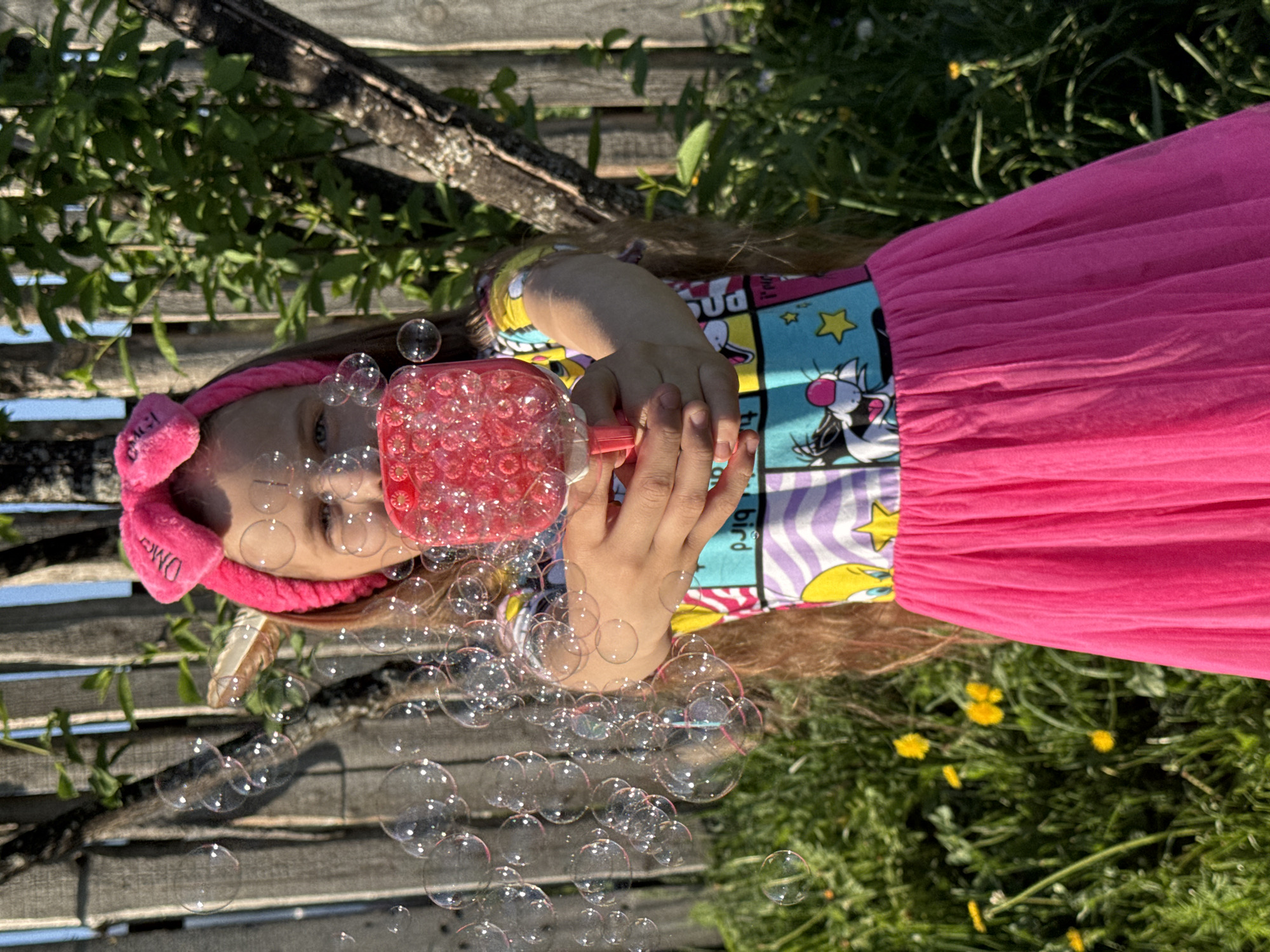 A young girl in Rusalevka, Russia, dressed in a colorful shirt and pink skirt, is blowing bubbles. She is wearing a pink headband. The bubbles are floating in the air. The background includes a wooden fence, green grass, and some small yellow flowers.