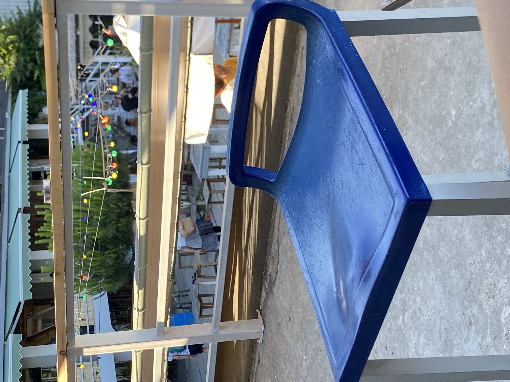 This media file shows a close-up of a blue plastic chair in the foreground, with a blurred background depicting an outdoor setting. The background appears to be a casual outdoor dining or gathering area, possibly a restaurant or bar with a rustic, open-air design. Several people are visible in the background, some sitting at tables and others standing, suggesting a social event or meal in progress. Strings of colorful lights are strung across the area, adding a festive atmosphere and hinting at it being evening or late afternoon. The overall impression is a relaxed, outdoor social gathering. The scene is outdoors, with greenery visible behind the structure, suggesting a warm weather environment. The lighting in the background suggests it is either late afternoon with long shadows cast, or early evening as the decorative lights are on. The context provided indicates the location is Varna, Bulgaria. There is no visible text in the image.