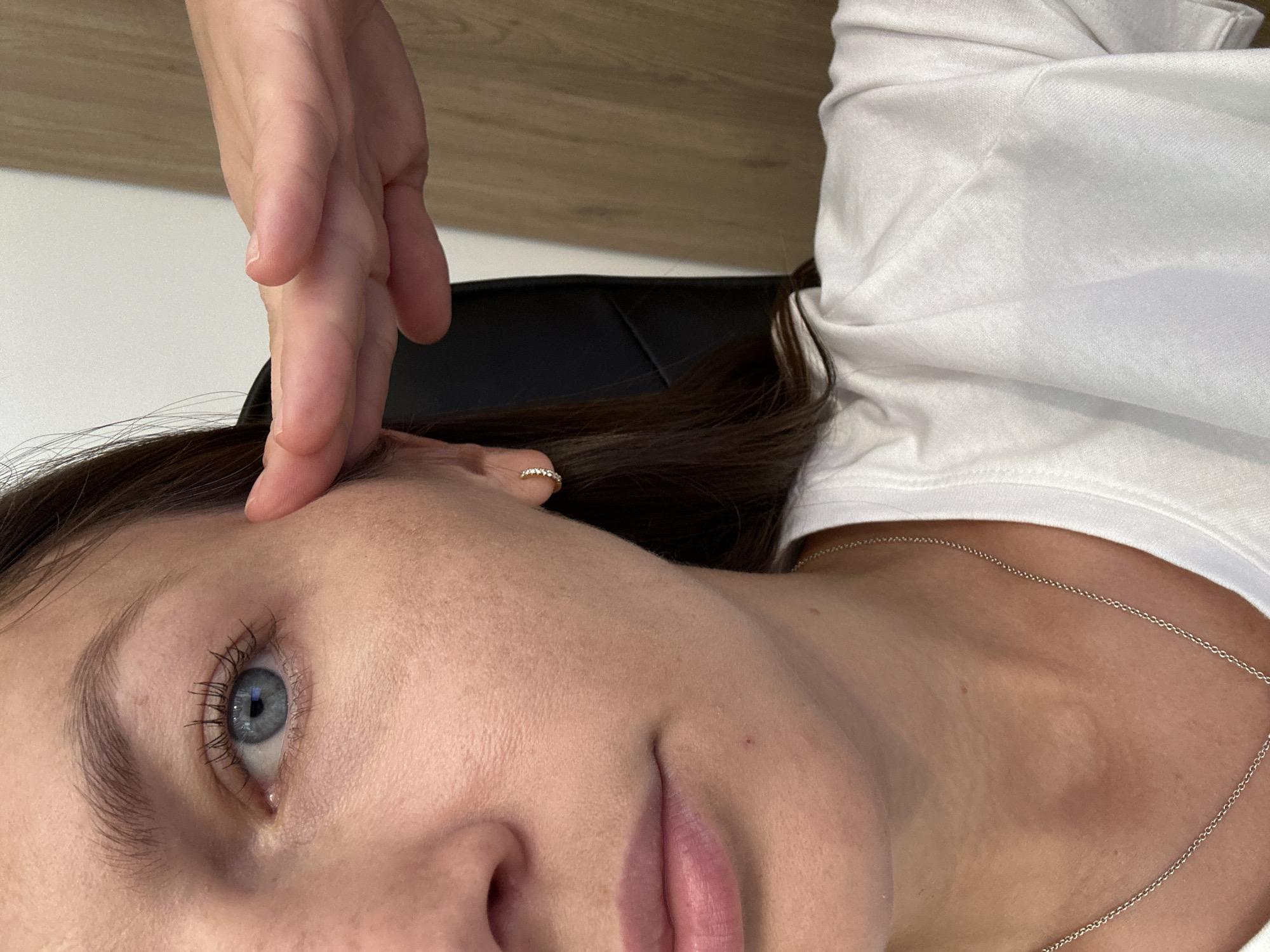A close-up, medium shot captures the right side of a person's face, focusing on their eye, cheek, mouth, and ear, with a hand obscuring part of their forehead and temple. The individual has fair skin, light-colored eyes with visible lashes, and dark brown hair. They are wearing a white t-shirt and a silver necklace. A small, gold hoop earring with tiny gems is visible on their right ear. The background consists of a dark chair and a wooden surface. The image was taken in Bečići, Montenegro.