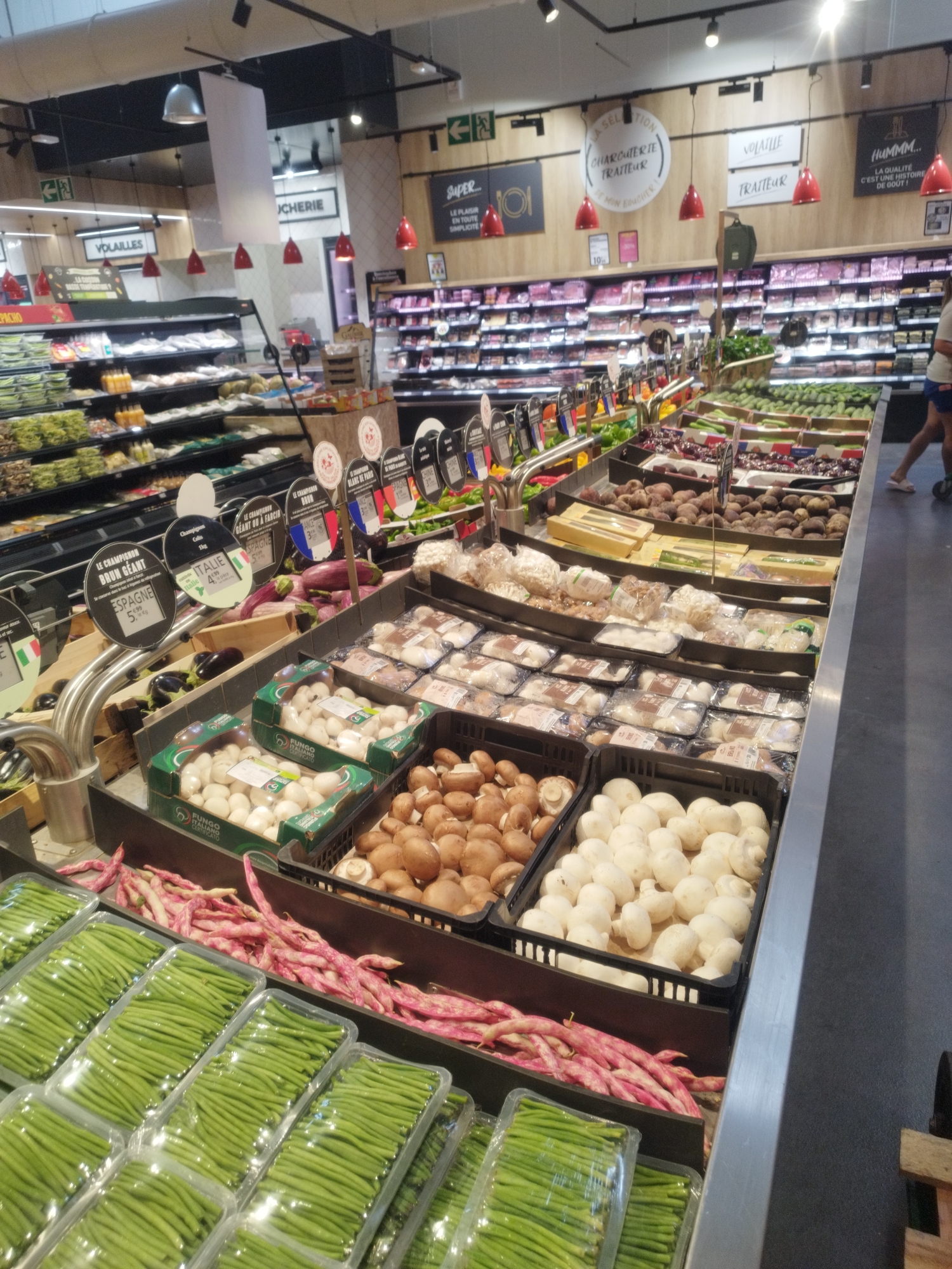 The image depicts the interior of a supermarket in Vallauris, France, specifically the produce section. The aisle is filled with various fresh vegetables displayed in refrigerated cases and baskets. In the foreground, rows of packaged green beans are visible. Behind them, a pile of pink beans is loosely arranged in a display. Further back, boxes of white mushrooms are prominently featured, alongside brown mushrooms. Other vegetables such as eggplants and beets are also present. The background reveals shelves stocked with packaged goods and signs indicating different product categories and origins, such as "VOLAILLES" (poultry), "TRAITEUR" (caterer), and "LA SELECTION CHARCUTERIE TRAITEUR". Red pendant lights hang from the ceiling, illuminating the displays. A person, partially visible on the right side of the image, is walking away from the produce section. The overall environment suggests a well-organized and brightly lit retail space focused on fresh produce.