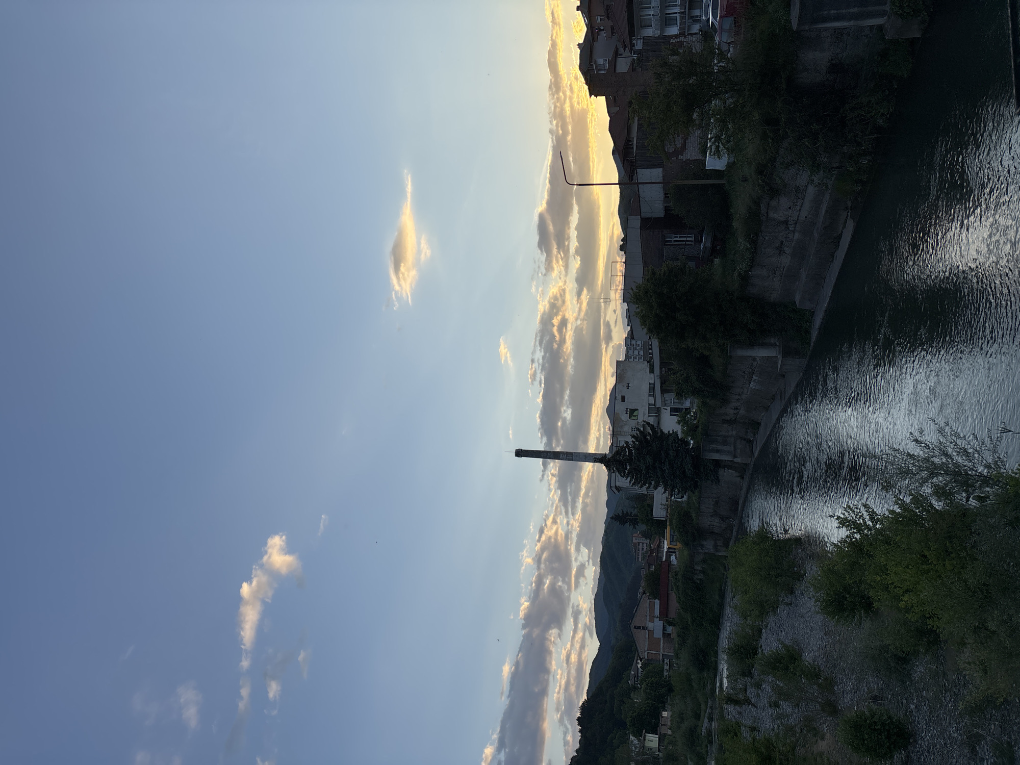 The image shows a view of Zlatograd, Bulgaria at dusk. A river or canal runs vertically through the right side of the frame, with concrete embankments and vegetation lining the banks. Buildings are visible on the embankment, including houses and apartment buildings. A tall smokestack rises in the center of the image, silhouetted against the sky. The sky is partially cloudy, with a clear blue section on the left and a band of golden-tinged clouds near the horizon. Hills and mountains are visible in the background. A street lamp stands on the right side of the frame along with the buildings.
