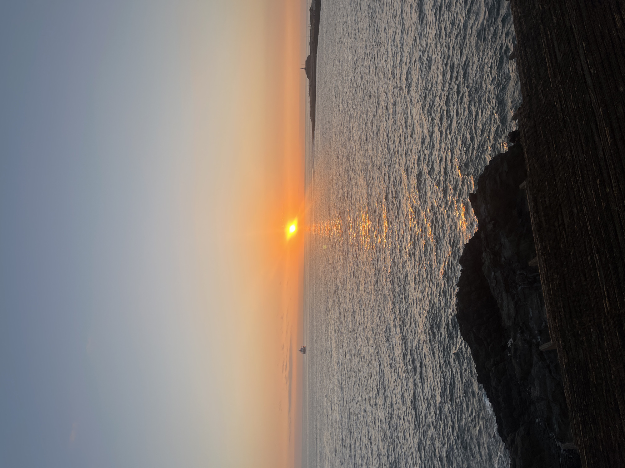 The image depicts a sunset over the ocean near Arica, Chile. The sun is setting on the horizon, casting a golden light on the water. To the right of the image, there's a rocky cliff face and wooden structure, suggesting a pier or coastal fortification. An oil rig is seen on the horizon. The sky is a gradient of colors, from light blue to orange near the sun.