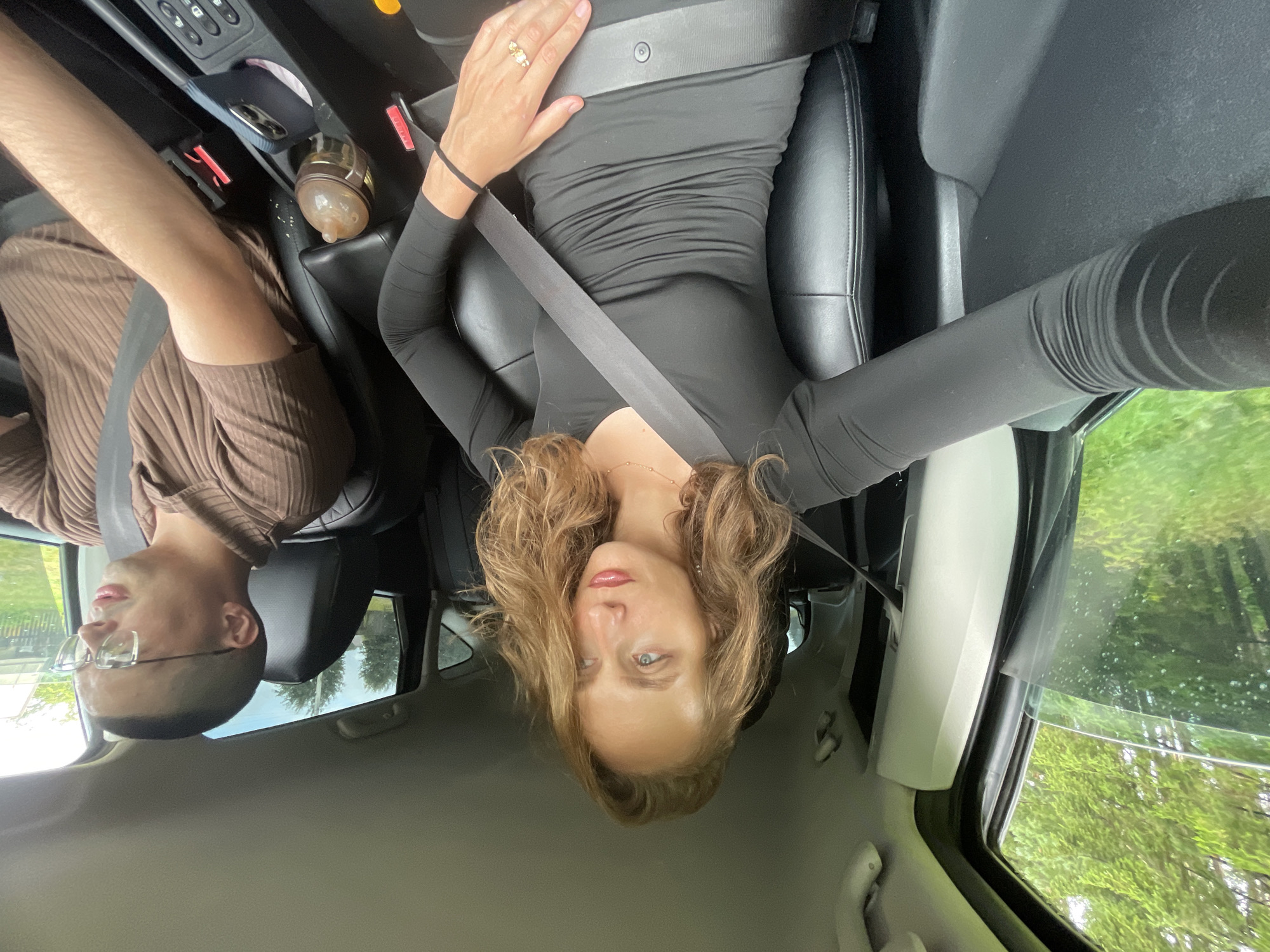 A woman with blonde, wavy hair is in the driver's seat of a car, wearing a black, long-sleeved dress and a seatbelt. She is looking to her right. A man with short hair and glasses, wearing a brown collared shirt, is in the passenger seat, also wearing a seatbelt. He is looking forward and to his right. The interior of the car is visible, including the dashboard and seats. Outside the car windows, green trees and foliage can be seen, suggesting a rural or suburban environment. The image was taken in Podshivalovo, Russia.