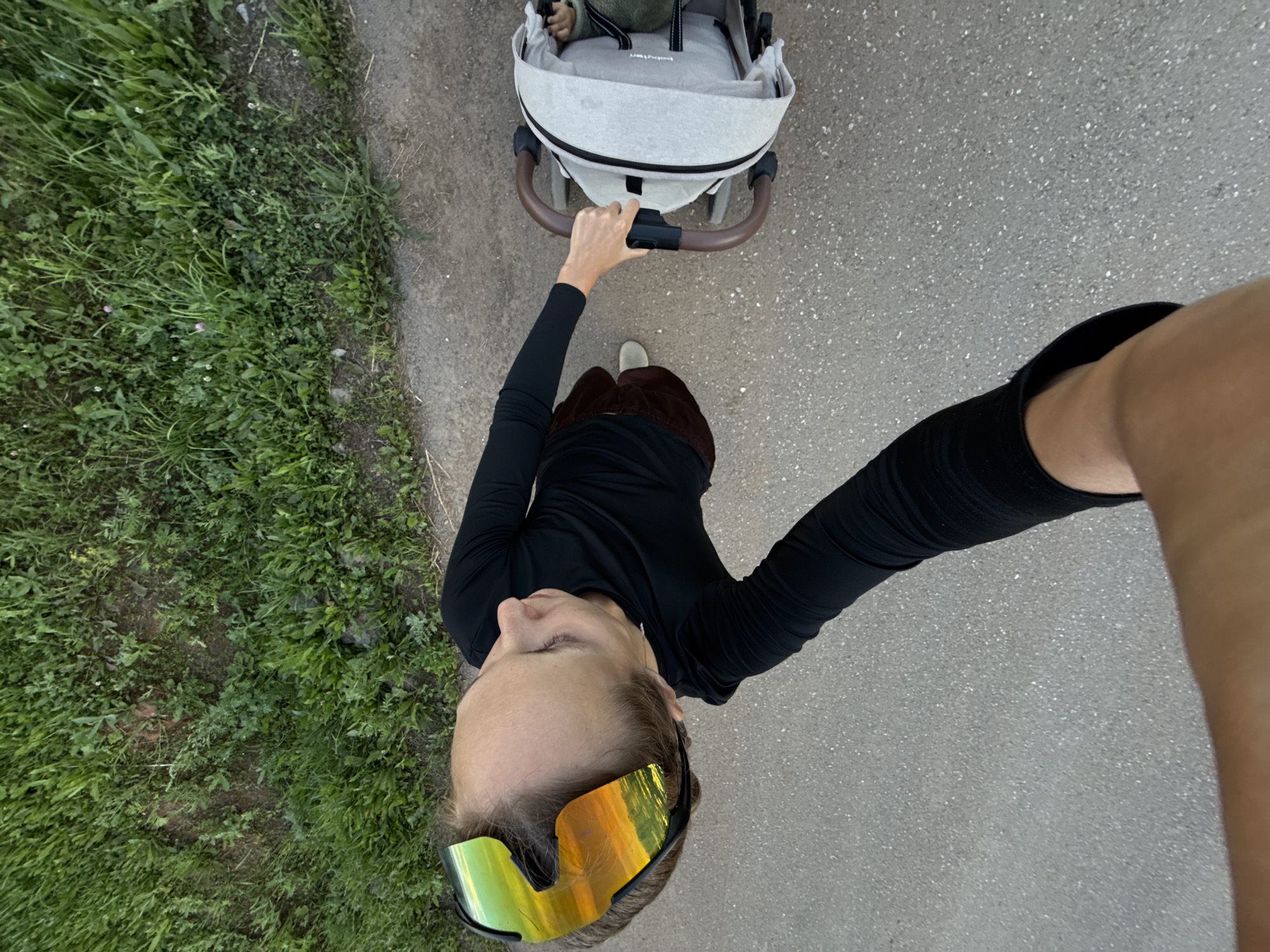 A person wearing ski goggles and a black long-sleeved shirt pushes a light grey baby stroller with a brown handle on a paved path. The person's right hand is on the stroller handle. To the left of the path is a grassy area with green grass and some dirt. The image is taken from a high angle looking down. The scene is likely outdoors in a park or along a sidewalk. The location is specified as Podshivalovo, Russia.