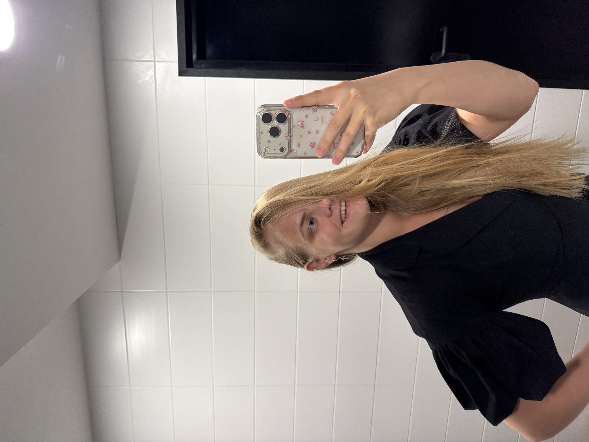 A young woman with long blonde hair is taking a selfie in a bathroom. She is holding an iPhone with a floral case. She is wearing a black top with puffy sleeves. The bathroom has white tiled walls and a black framed mirror. There is no evidence to suggest the photo was taken in Izhevsk, Russia.