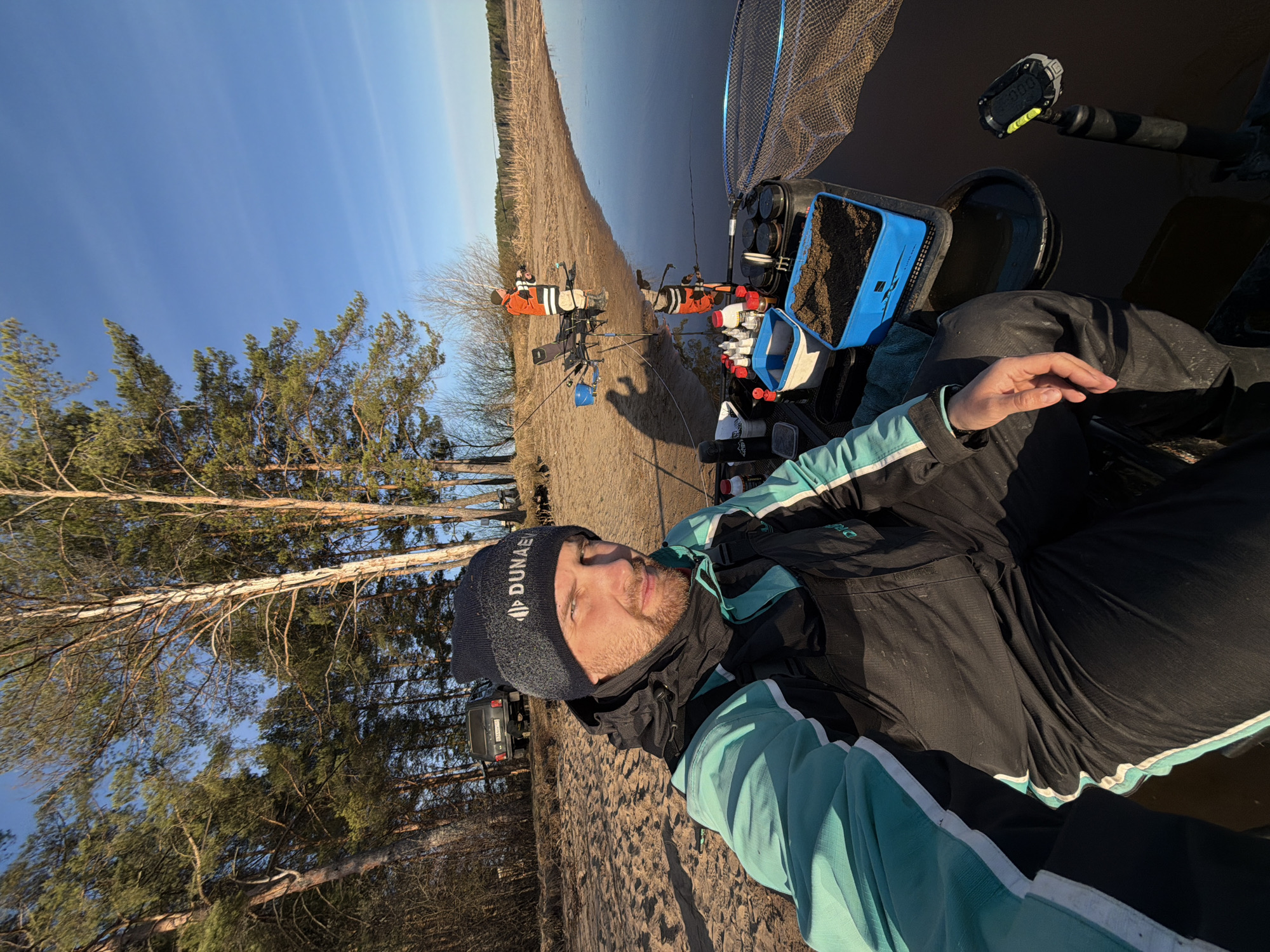 This media file presents a first-person, selfie-style photograph of a man fishing in an outdoor, natural setting near Chaykovsky, Russia. The man, the primary subject, is in the foreground, looking directly at the camera. He has a beard and is wearing a dark beanie with the text "DUNAEV" in white, a black and teal/mint green jacket, and dark pants. His right hand is visible in the lower right portion of the frame.
To the man's right, an assortment of fishing gear is meticulously arranged. This includes a large fishing net, a blue container filled with brown granular bait, and various smaller containers and bottles. A digital device, possibly a timer or scale, displaying "0.00" is also attached to some equipment.
The background reveals a serene landscape. A wide, tranquil body of water, likely a river or lake, extends across the scene to the horizon, reflecting the clear blue sky. A sandy or dirt bank separates the water from a wooded area. Further along the bank, two other individuals are visible, also engaged in fishing with their rods set up on stands; they are wearing bright orange and white outerwear.
Tall pine trees and some bare-branched deciduous trees populate the left side and background of the image. A dark-colored SUV is partially visible among the trees in the far background. The sky is a clear, bright blue, indicating sunny weather, and the warm light suggests either morning or late afternoon. The scene depicts a calm and focused fishing activity in a picturesque, natural environment.