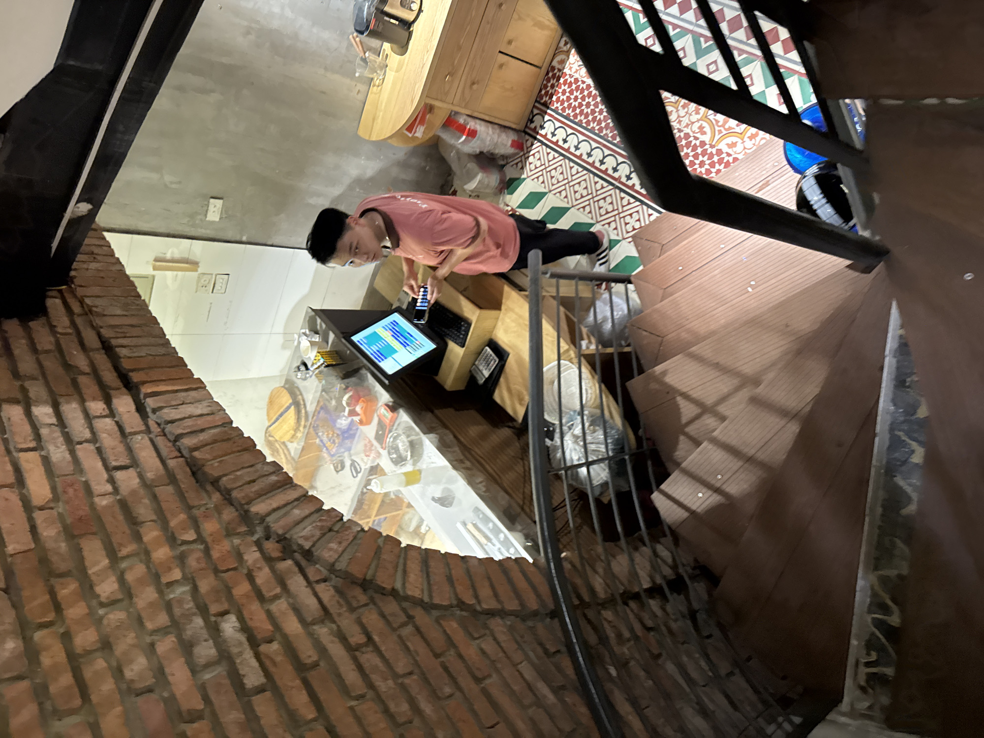 A young Vietnamese male, wearing a pink t-shirt and black pants, stands at a counter with a computer monitor and calculator in what appears to be a cafe or restaurant in Hanoi, Vietnam. The male is looking at a mobile phone. The environment includes a spiral wooden staircase with metal railings in the foreground. The walls are made of exposed brick and concrete. The floor is tiled with a colorful, geometric pattern. A wooden cabinet with drawers is visible to the right of the counter.
