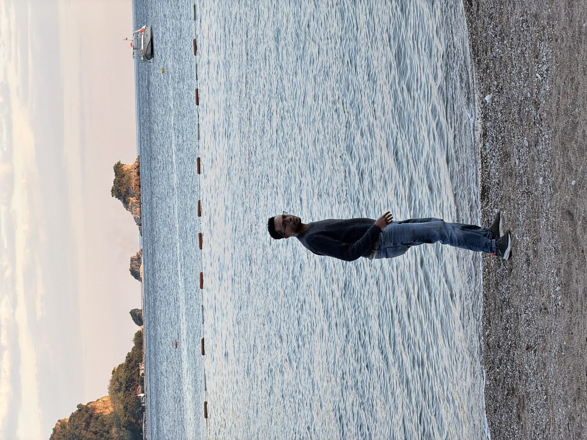 A man stands on the gravelly shore of a body of water in Budva, Montenegro. The water, likely the Adriatic Sea, stretches out toward a coastline with rocky outcrops and greenery. A boat is visible in the distance. The man is wearing jeans, a dark sweater, and sneakers. The sky is bright, suggesting either early morning or late afternoon. A line of buoys floats along the water.
