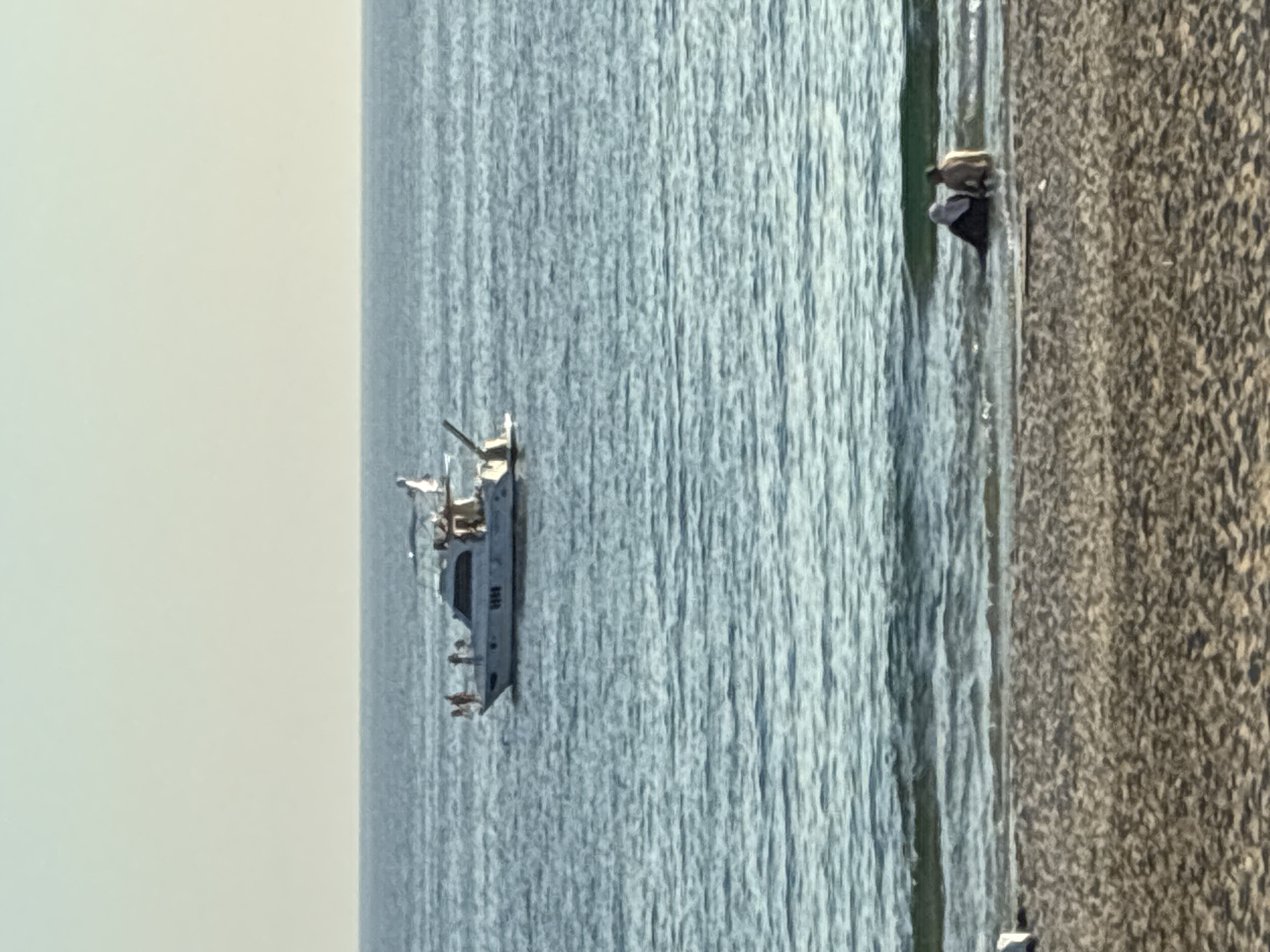 A distant view of the sea and beach in Alanya, Turkey. On the water, a white motor yacht with several people on board is visible. Near the shore, two individuals, one wearing a headscarf, sit partially submerged in the shallow water. The beach in the foreground is sandy. The sky is clear and pale.