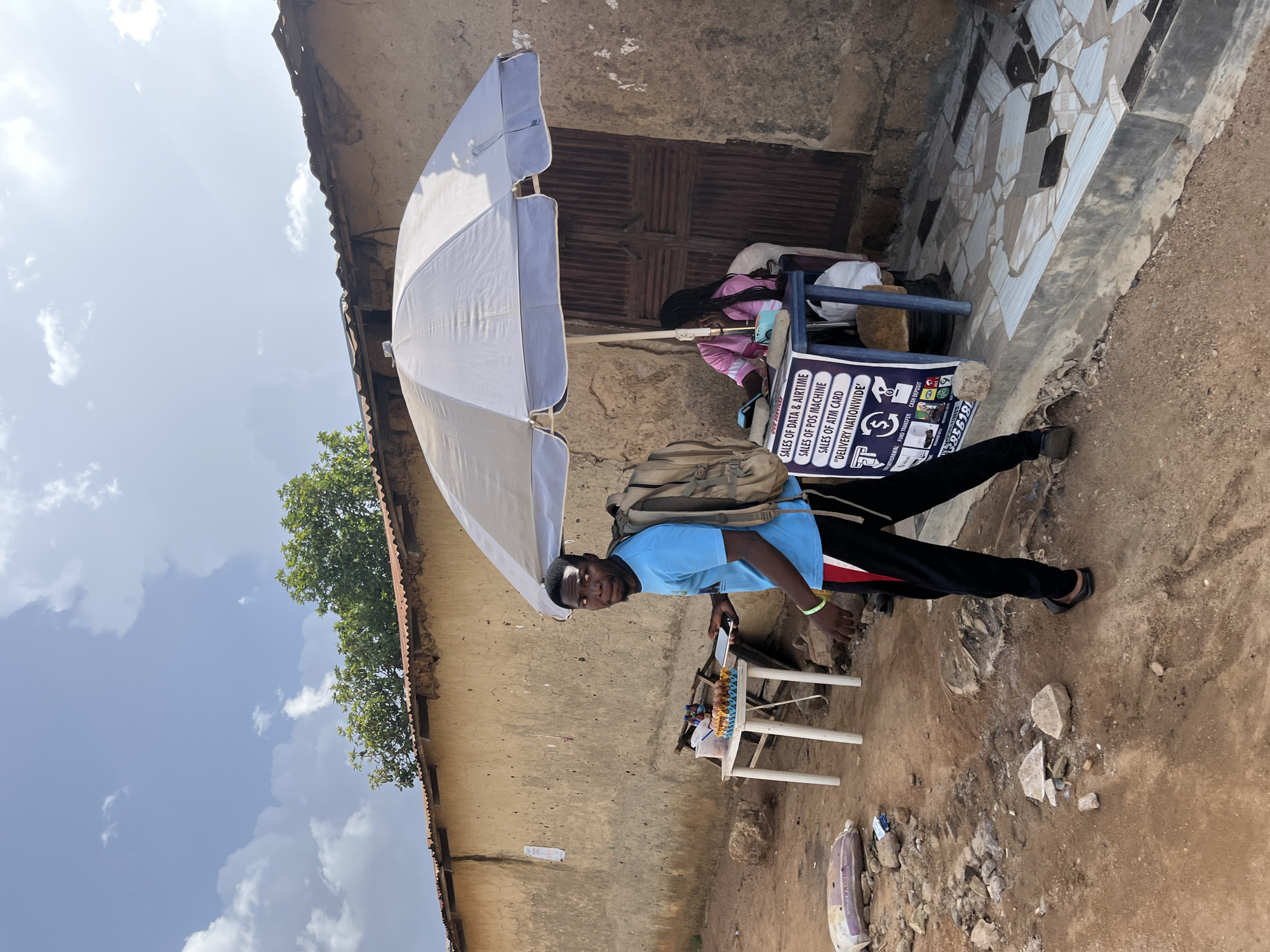 A young man wearing a blue t-shirt, black pants with red and white stripes, and a backpack stands on a dirt road in front of a building in Jos, Nigeria. He is holding a tablet and looking towards the camera. To his right, a woman in a pink shirt sits under a large white umbrella, attending to a stall. The stall displays a sign listing services such as "SALES OF DATA & AIRTIME," "SALES OF POS MACHINE," "SALES OF ATM CARD," and "'DELIVERY NATIONWIDE'." A small white table to the left of the stall holds a tray of eggs and bottles of liquid. The building behind the stall appears to be made of mud with a corrugated metal roof. The ground is a mix of dirt, rocks, and tiled pavement. The sky is partly cloudy.