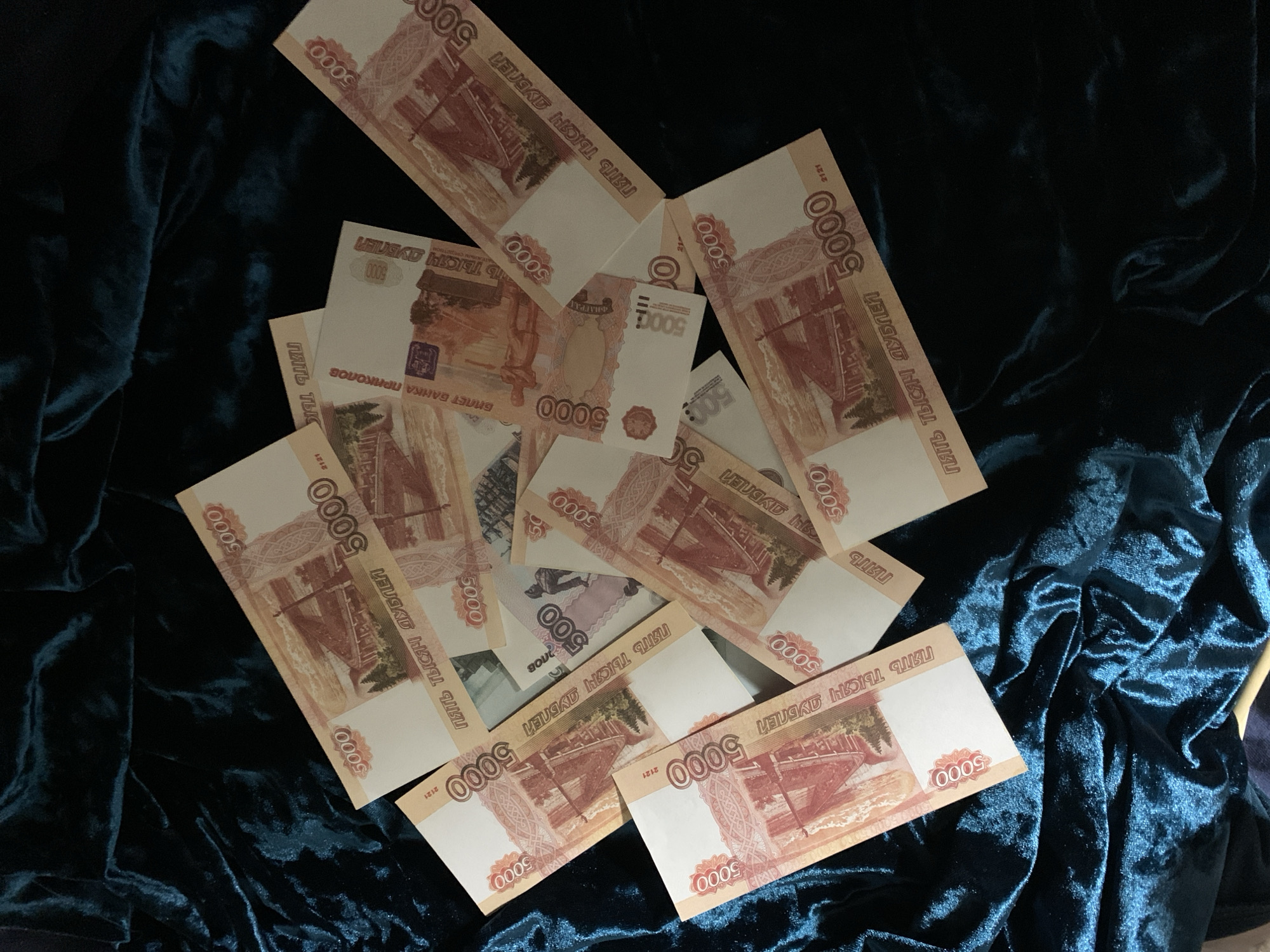 A collection of 5000 Russian ruble banknotes are scattered on a dark blue velvet fabric surface. The banknotes feature the number "5000" prominently and depictions of a landscape. There are multiple banknotes overlapping each other. There are a few smaller denomination notes amongst the larger ones. Some of the notes are labeled as "bank notes of fun". The background is completely filled by the blue velvet fabric.