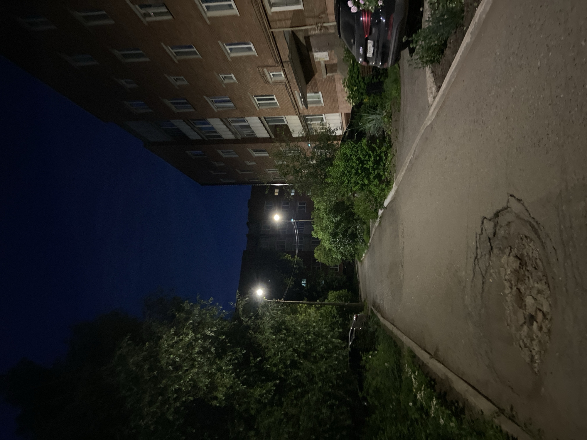 This media file depicts an outdoor scene at night in Votkinsk, Russia. The primary subjects are a multi-story brick apartment building, a paved walkway with a broken section, lush green trees and bushes, and a dark-colored car parked partially obscured by vegetation. The apartment building has many windows, some of which are illuminated. Streetlights are visible, casting a warm glow on the surroundings.
The scene appears to be a quiet residential area. The time of day is clearly night, indicated by the dark sky and the artificial lighting. The weather appears to be clear, with no visible precipitation. The broken section of the pavement suggests a potentially worn or aged infrastructure.
There are no people visible in the image. The overall mood is serene and perhaps a little melancholic due to the darkness and the state of the pavement. The presence of trees and greenery suggests a possible summer or late spring season. The license plate of the car is partially visible but the text is illegible.