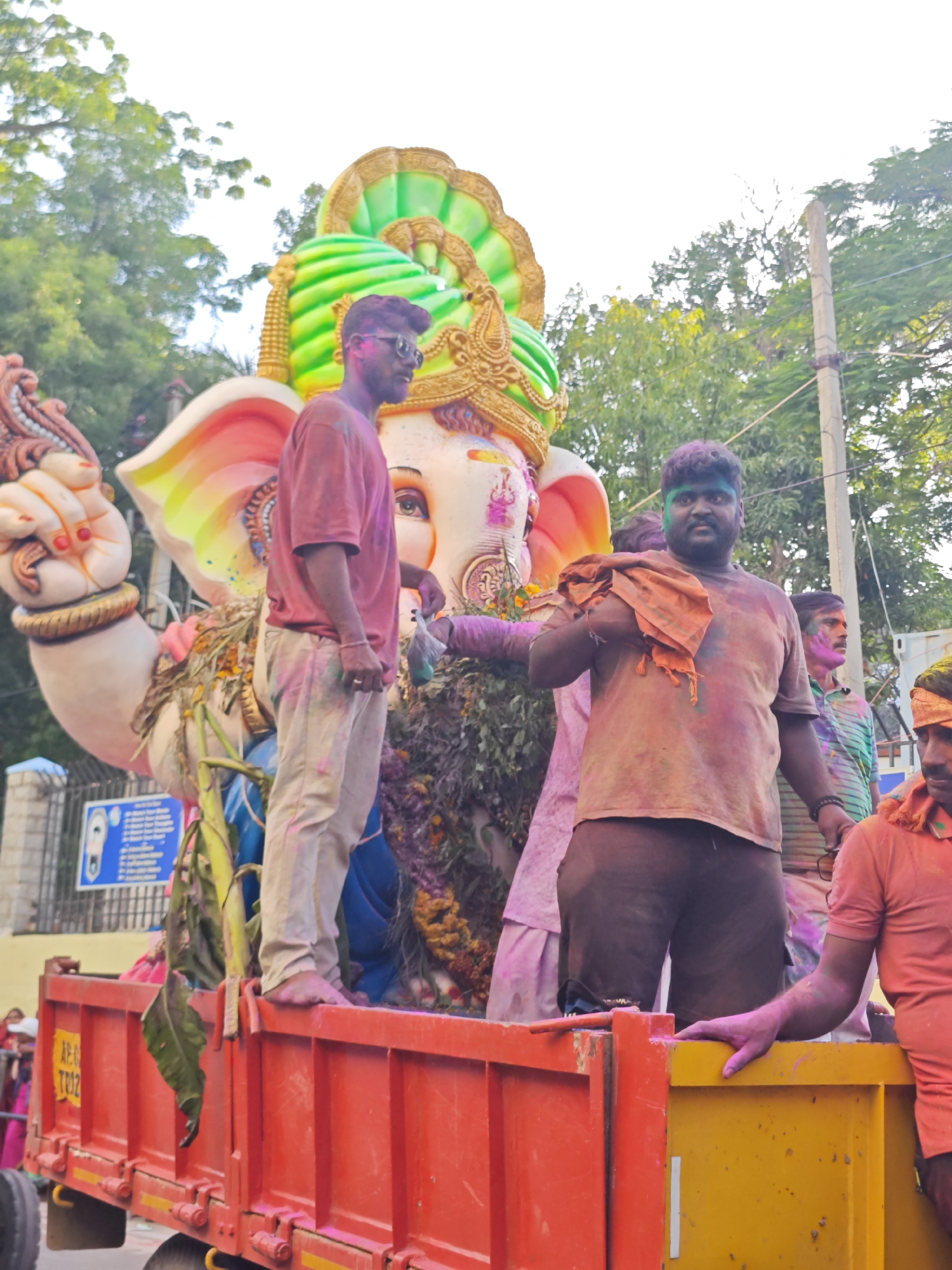 In the city of Puttaparthi, India, on a bright, overcast day, a colorful Ganesh idol is being transported on the back of a red flatbed truck. The idol, adorned with green and gold ornaments, is massive, with its trunk and large ears prominently visible. Several men are standing around the idol, covered in vibrant Holi powder, suggesting a festival or celebration is underway. The man in the foreground, to the right of the idol, wears a light brown t-shirt and dark pants, his face and body smudged with pink and orange powder. He holds an orange cloth draped over his shoulder. To his left, another man, wearing a maroon t-shirt and light-colored pants, stands with his left arm raised, a plastic bag in his hand. His face is partially obscured, but sunglasses are visible. Behind them, other individuals are partially seen, also dusted with Holi colors. The truck is decorated with foliage and flowers around the idol. In the background, lush green trees and a utility pole with wires line the street, and a blue sign with text is visible on a fence. The overall scene depicts a lively procession, characteristic of the Ganesh Chaturthi festival.