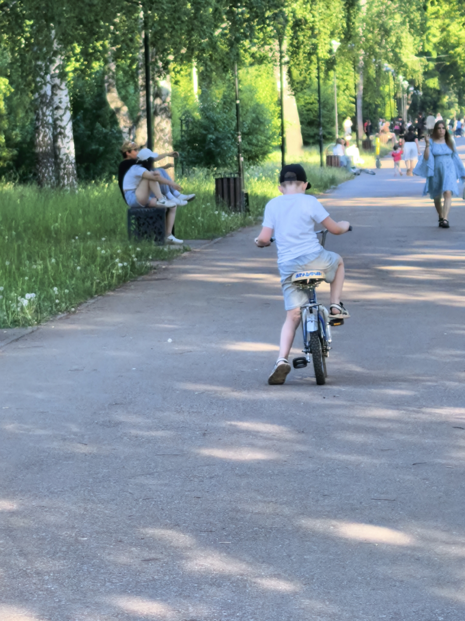 A boy rides a bicycle on a paved path in a park. He is wearing a white t-shirt, denim shorts, sandals, and a black baseball cap. Two people are sitting on a park bench on the left. A woman in a light blue dress walks on the path ahead of the boy. Other people can be seen in the background. Trees line the path. The location is Sterlitamak, Russia.