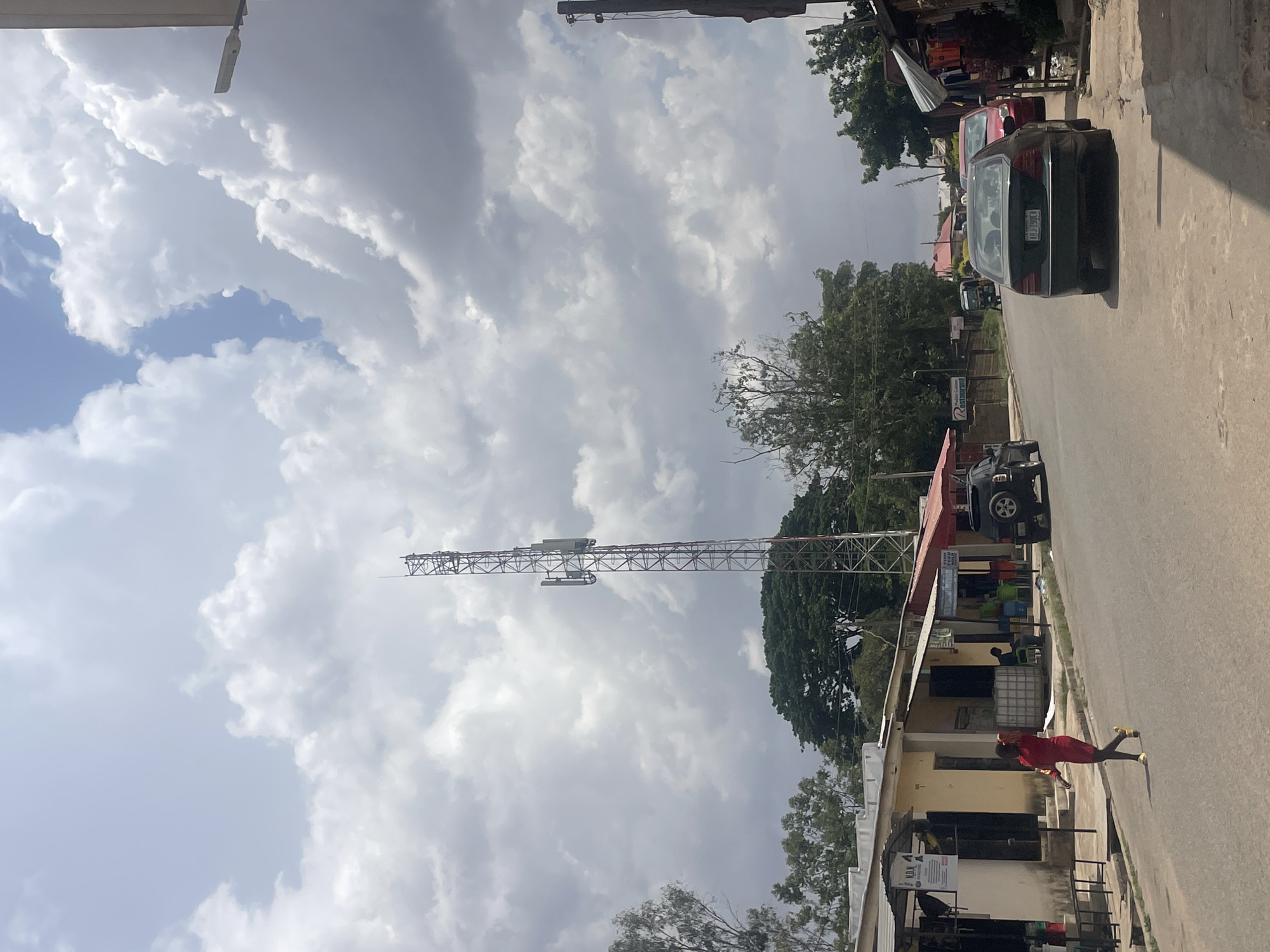 The image shows a street scene in Jos, Nigeria. A tall telecommunications tower rises in the background against a cloudy sky. The street is lined with low buildings, some with awnings and businesses. Several vehicles are parked on the side of the road, including a dark-colored car in the foreground. A woman wearing a red dress and yellow shoes is walking on the sidewalk. The environment is a mix of urban and natural elements, with trees interspersed among the buildings.