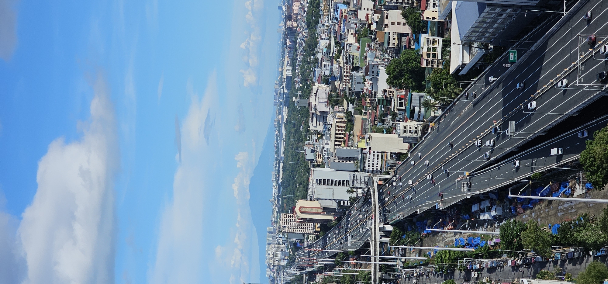 The image shows an aerial view of a multi-lane highway in Makati City, Philippines, under a bright blue sky with scattered clouds. The highway is elevated and curves through a dense urban landscape filled with buildings of varying heights, from low-rise structures to skyscrapers. Numerous vehicles are visible on the highway, indicating moderate traffic flow. To the left of the highway, there is an area that appears to be under construction or development, with blue tarpaulins covering materials and some small structures. Trees and greenery are interspersed throughout the cityscape, particularly along the sides of the highway and between buildings. In the background, a mountain range is faintly visible on the horizon, suggesting a broader geographical context beyond the immediate urban sprawl. The scene captures a typical day in a bustling metropolitan area.