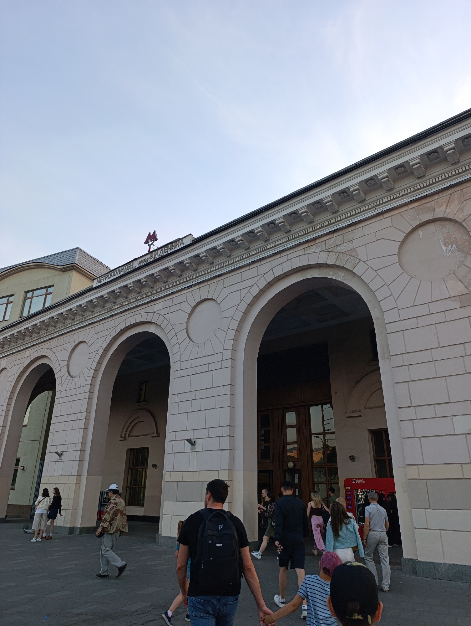 This image depicts the exterior of a grand, classical-style building, identified as an entrance to the Moscow Metro, specifically likely the Kurskaya station (Koltsevaya Line) in Moscow, Russia, under a clear, light blue sky.
The building's facade is constructed from light-colored stone or stucco, featuring a series of at least three large, deeply recessed arched openings that serve as entrances. Above these arches, a prominent cornice with decorative corbels runs horizontally across the building. A red neon sign is affixed to the building above the central arches, displaying the Cyrillic text "МЕТРОПОЛИТЕН имени ЛЕНИНА" (METROPOLITEN imeni LENINA), with the iconic red "M" logo of the Moscow Metro positioned centrally above the text. The building exhibits some signs of age and wear, particularly discoloration on the upper facade. To the left, an adjacent building section with a dark-tiled or slate roof and rectangular windows is visible.
A paved pedestrian area, likely dark grey flagstones or cobblestones, extends in front of the building. Approximately 15-20 individuals are present on this walkway, engaged in various actions. In the immediate foreground, a man wearing a black t-shirt, blue jeans, and a black backpack is seen from behind, holding the hand of a child wearing a striped shirt and a dark cap. Both are walking towards the metro entrance. To their right, a man in a light grey shirt and light trousers walks into one of the arched entrances. Further right, a woman in a light blue cardigan and white skirt is also approaching the entrance.
In the mid-ground, a man in a camouflage jacket and a white baseball cap, wearing grey trousers, walks towards the left side of the frame. Near the leftmost archway, two women are visible; one in a light-colored top and shorts, the other in a light top and skirt, appearing to be standing or conversing. Several other people, mostly adults, are walking into or out of the main entrances, suggesting a busy public thoroughfare. Near one of the rightmost entrances, a red automated kiosk or vending machine, possibly an ATM, is visible. The overall scene suggests an active urban environment during daylight hours.