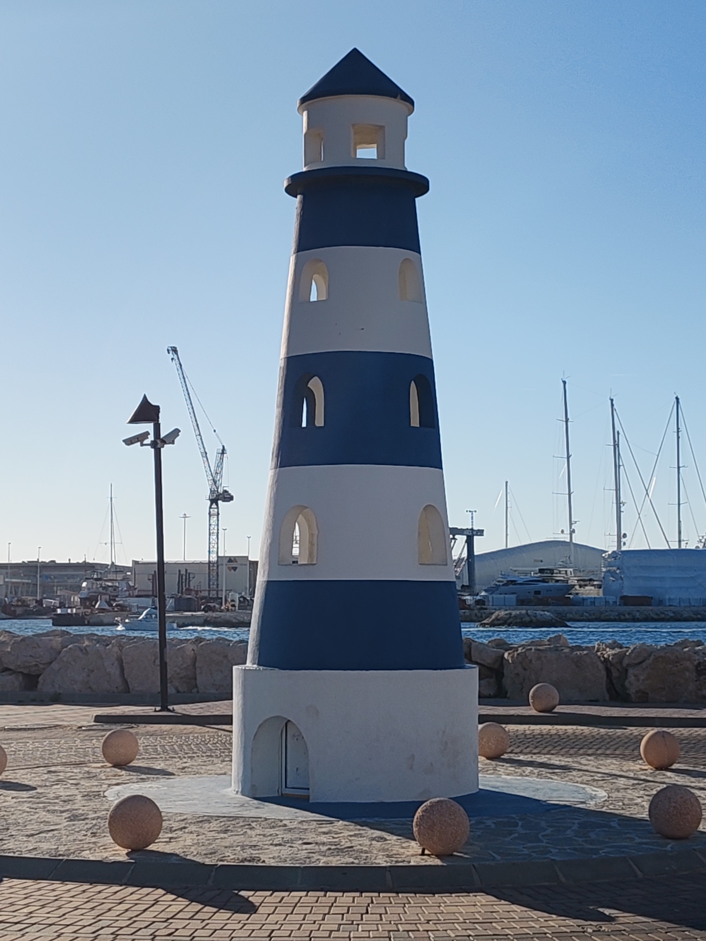 The image shows a striped, blue and white decorative lighthouse on a paved area in Dénia, Spain. The lighthouse has several arched windows and a small dark blue roof. The base has a small arched door. The lighthouse is surrounded by stone spheres, which are sitting on a paved area made of irregularly shaped stone tiles, bordered by red brick pavement. The sea is visible in the background, with boats, cranes, and buildings visible in the distance. A modern light post is visible to the left of the lighthouse. The sky is clear and blue.