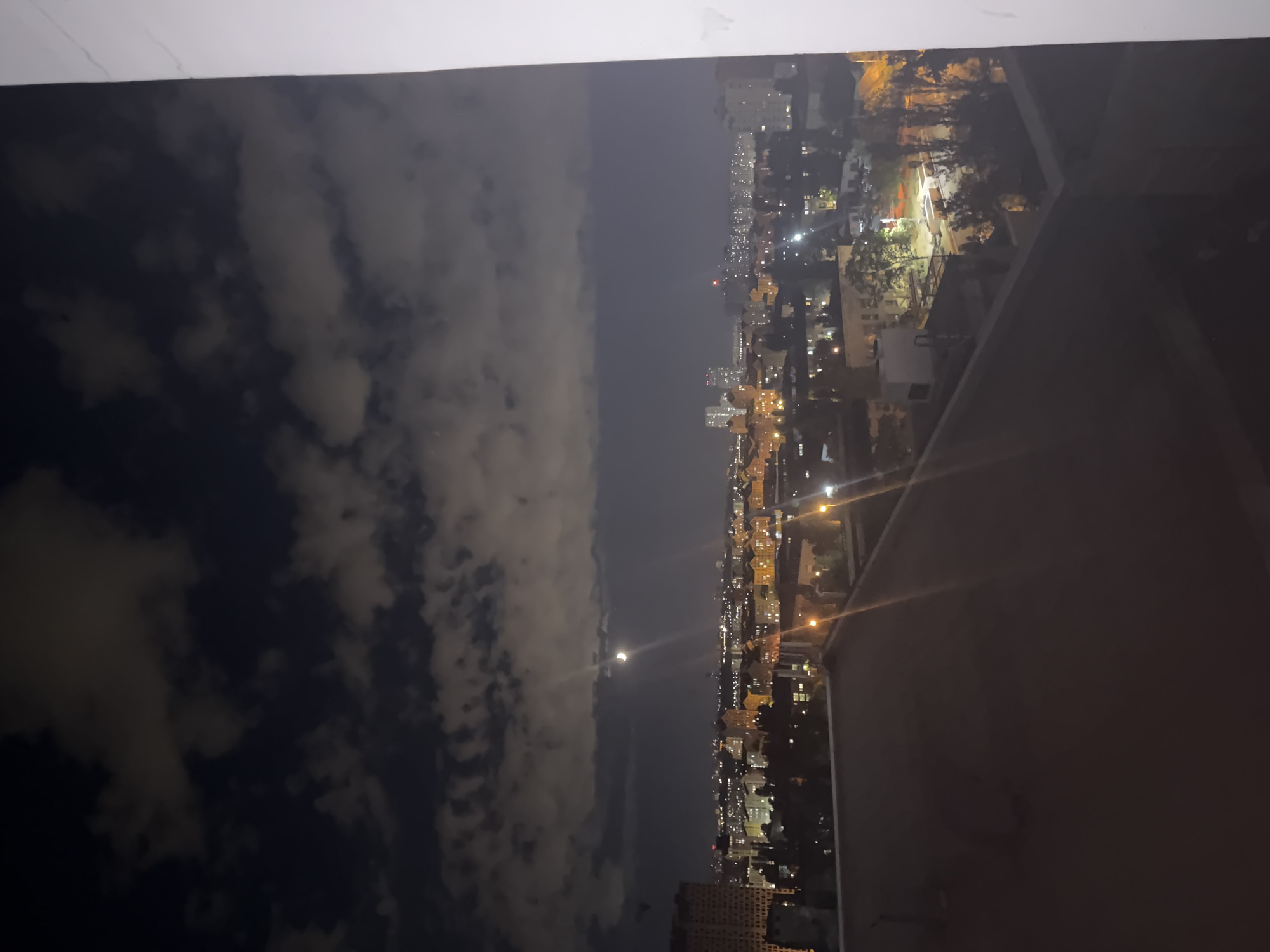 The image captures a nighttime view of a cityscape, presumably Kyiv, Ukraine, as indicated by the hint. The sky is dark, with scattered clouds illuminated by the moon, which is visible as a crescent. The city below is dotted with lights from buildings, suggesting an urban environment. In the foreground, a rooftop or elevated structure is visible, casting shadows. The composition is dominated by the night sky and the illuminated city, creating a serene, albeit slightly moody, atmosphere. There are no people or specific actions depicted in the scene. The overall impression is that of an urban landscape at night under a cloudy moonlit sky.
