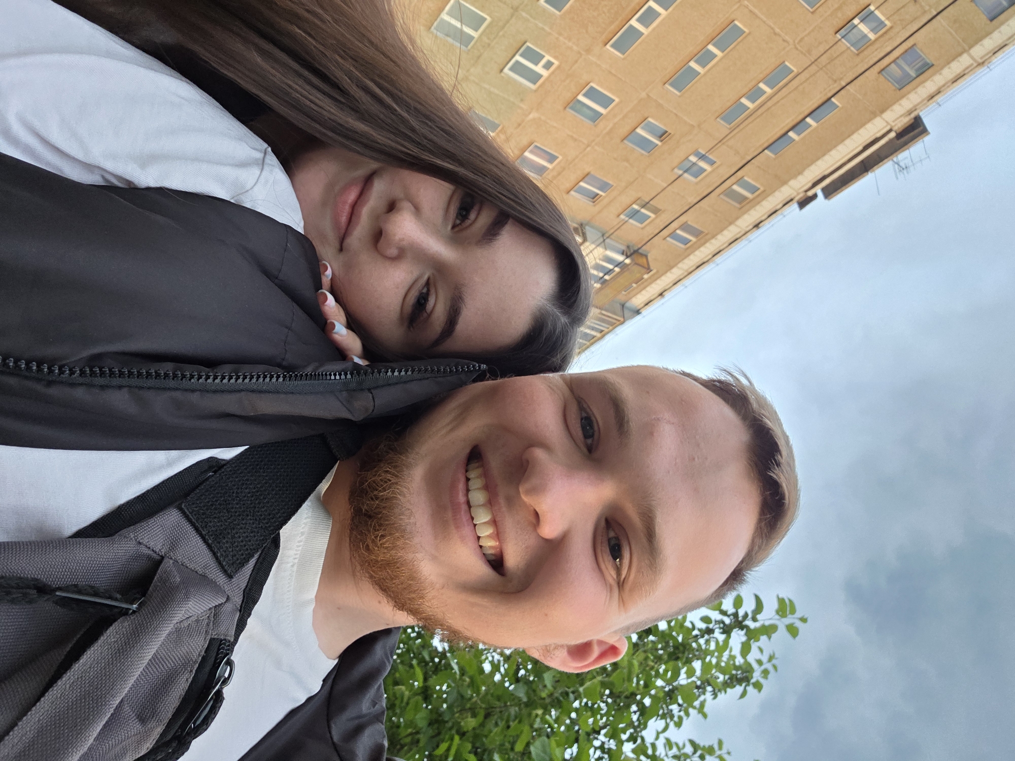 A selfie shows a man and a woman. The man has reddish hair and a beard, and he is smiling. The woman is behind him with long brown hair, and her face is partially visible. They are both wearing white shirts and black jackets. The man has a gray bag slung over his shoulder. Behind them, there is a building with rows of windows. The sky is visible, and it appears to be overcast. The photograph appears to have been taken in Perm, Russia, based on the architectural style of the building. The couple appears to be enjoying a stroll.