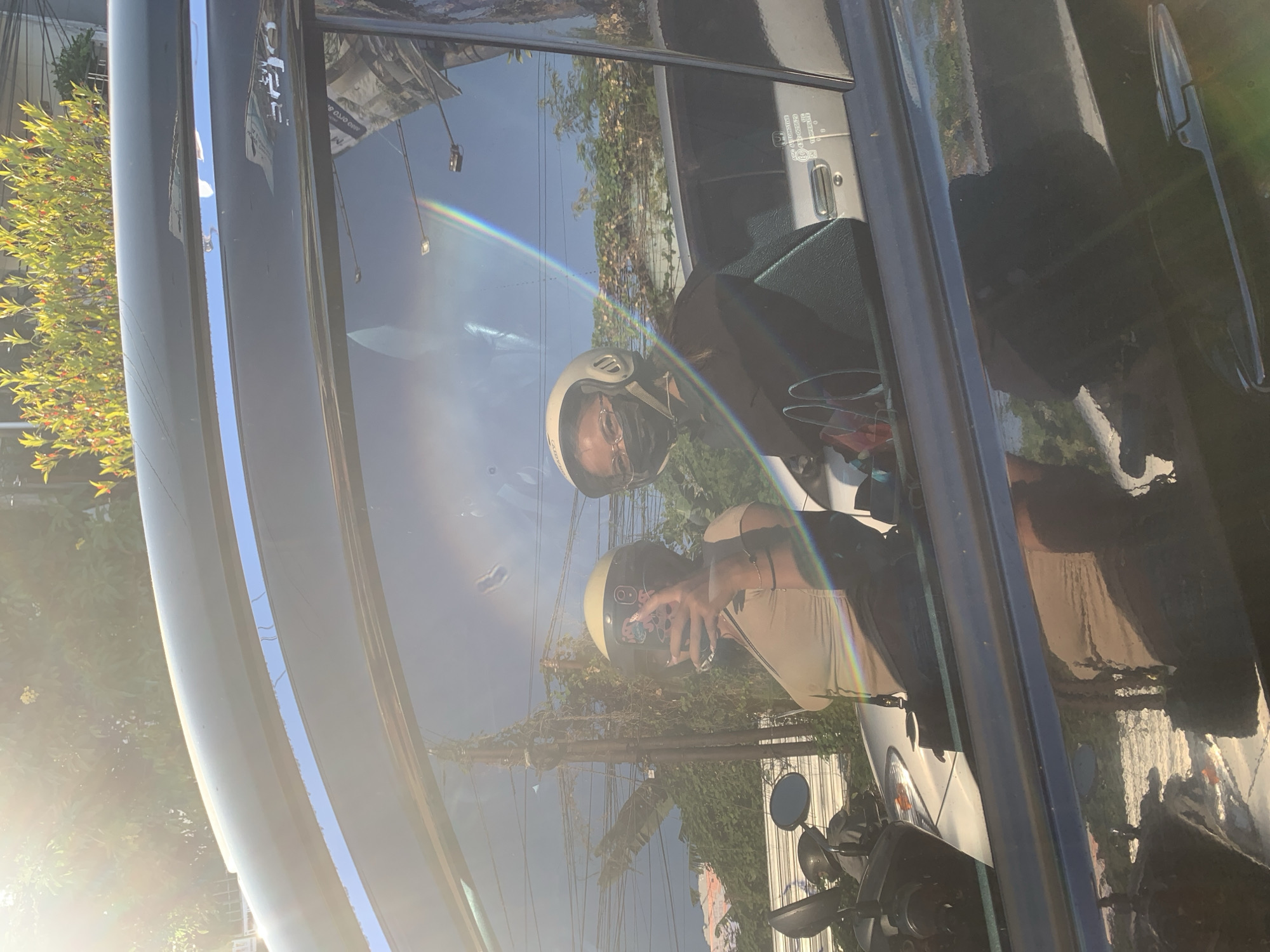 Two individuals, one in the foreground and one behind, are seated on a motorcycle. The individual in the foreground, appearing to be a woman, is wearing a tan tank top and a white helmet. She is holding a smartphone in her right hand and taking a selfie. The second individual, also appearing to be a woman, is wearing a dark shirt and a white helmet, with a black face mask covering her mouth and nose. Both are visible through the reflection in a car window. The background depicts a street scene with power lines and trees, consistent with the urban environment of Denpasar Barat, Indonesia, illuminated by bright sunlight. A rainbow-like lens flare is visible across the car window.