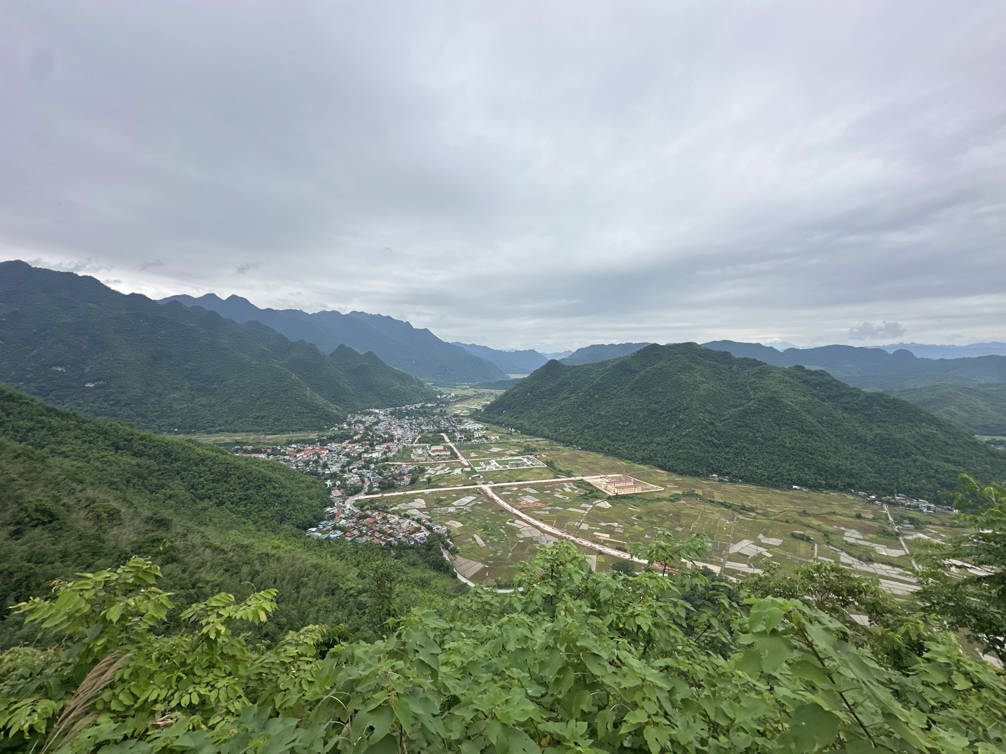 The image provides a panoramic view of a valley in Xóm Khán, Vietnam, under a cloudy sky. In the foreground, lush green foliage obscures the lower part of the frame. The midground reveals a sprawling town with numerous buildings, characterized by red-tiled roofs, clustered along a winding road. Beyond the town, expansive agricultural fields, some appearing to be flooded rice paddies reflecting the sky, dominate the valley floor. A main road with traffic infrastructure cuts through these fields, leading to more structured buildings, possibly educational or administrative complexes, some with yellow facades. The background is filled with rolling, densely forested green mountains that form the natural boundaries of the valley, their peaks fading into the hazy distance. The overall scene depicts a rural Vietnamese landscape where human settlement and agriculture coexist with a mountainous environment.