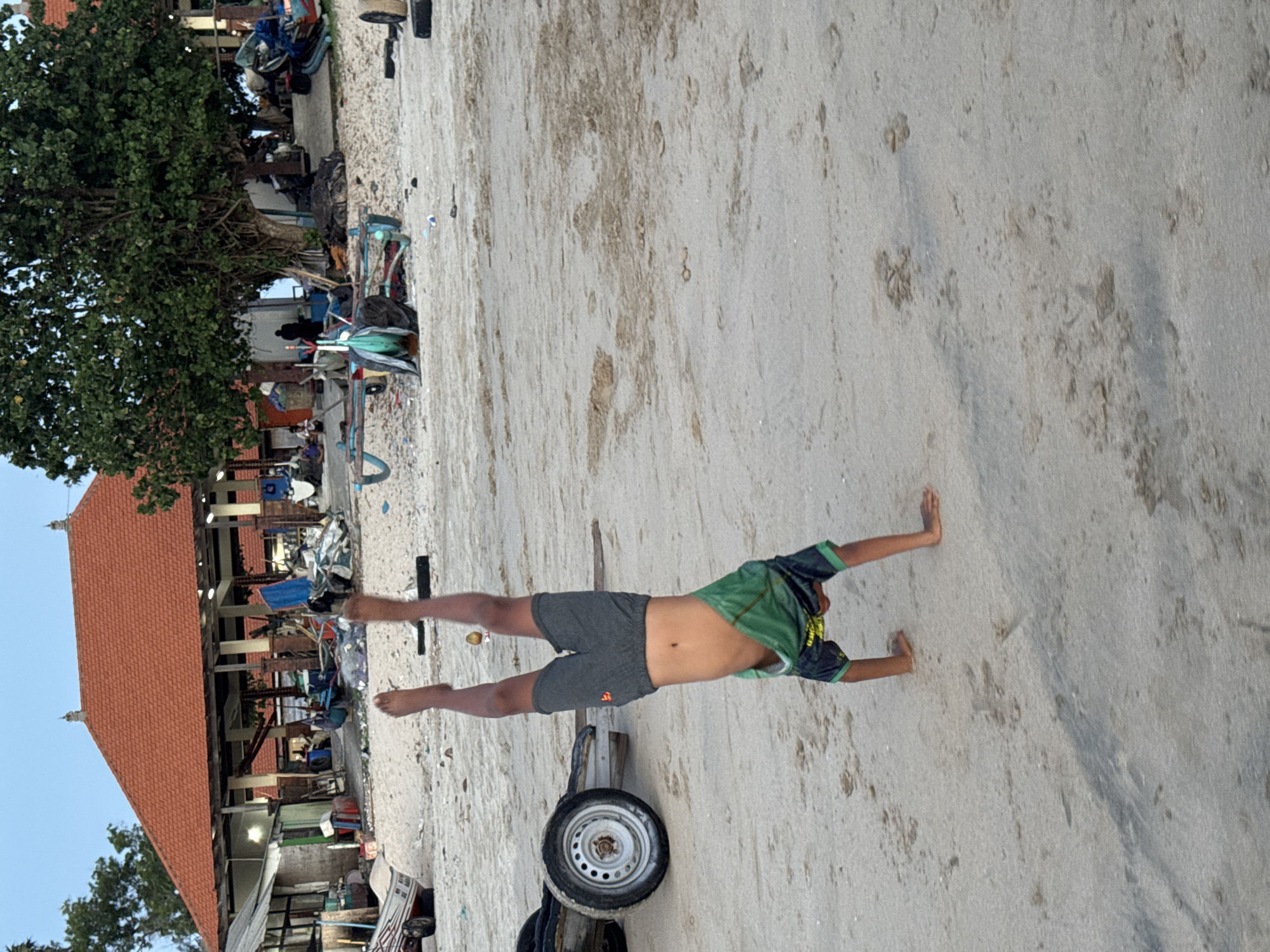A low-angle shot features a child lying prone on a sandy beach, with arms and legs extended. The child wears gray shorts and a green patterned shirt, and their bare abdomen is visible. Behind the child is a small trailer with a single wheel and wooden supports resting on the sand.
To the left of the child and trailer is a complex scene of buildings and trees. A red-tiled roofed building is partially visible, flanked by trees and utility poles. Structures and miscellaneous objects are clustered together, creating a cluttered, lived-in area. The location is Kuta Selatan, Indonesia.