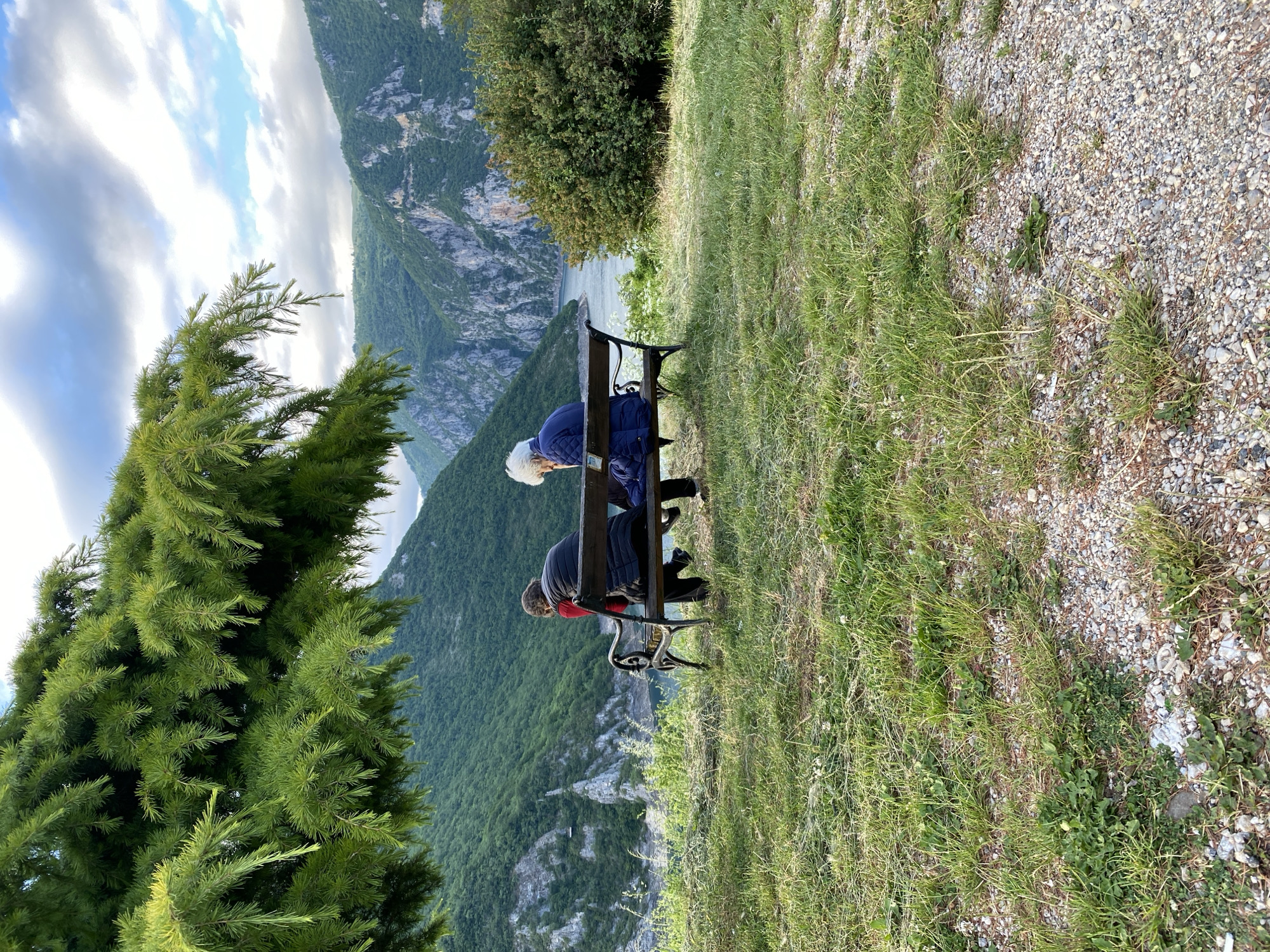 The image captures a picturesque landscape view from an elevated vantage point in Plužine, Montenegro. Two people are seen sitting on a dark wooden and metal park bench, with their backs to the camera, gazing out at the expansive natural scenery. The person on the left wears a blue quilted jacket and has white or blonde hair, while the person on the right is dressed in a dark jacket with a hint of red underneath.
They are observing a majestic vista of rugged, green-clad mountains that descend steeply into a valley, where a serene body of water, likely a lake or reservoir, is visible. The sky above is partly cloudy, featuring a mix of white and grey clouds against patches of blue, indicating it is daytime, possibly late morning or afternoon.
In the immediate foreground on the left, a vibrant green evergreen tree with dense needles partially frames the scene. The ground in the foreground consists of a mix of green grass and a gravel path or ground covering. The overall scene conveys a sense of peaceful contemplation and appreciation for the grand, natural beauty of the Montenegrin wilderness. No text is visible in the image.