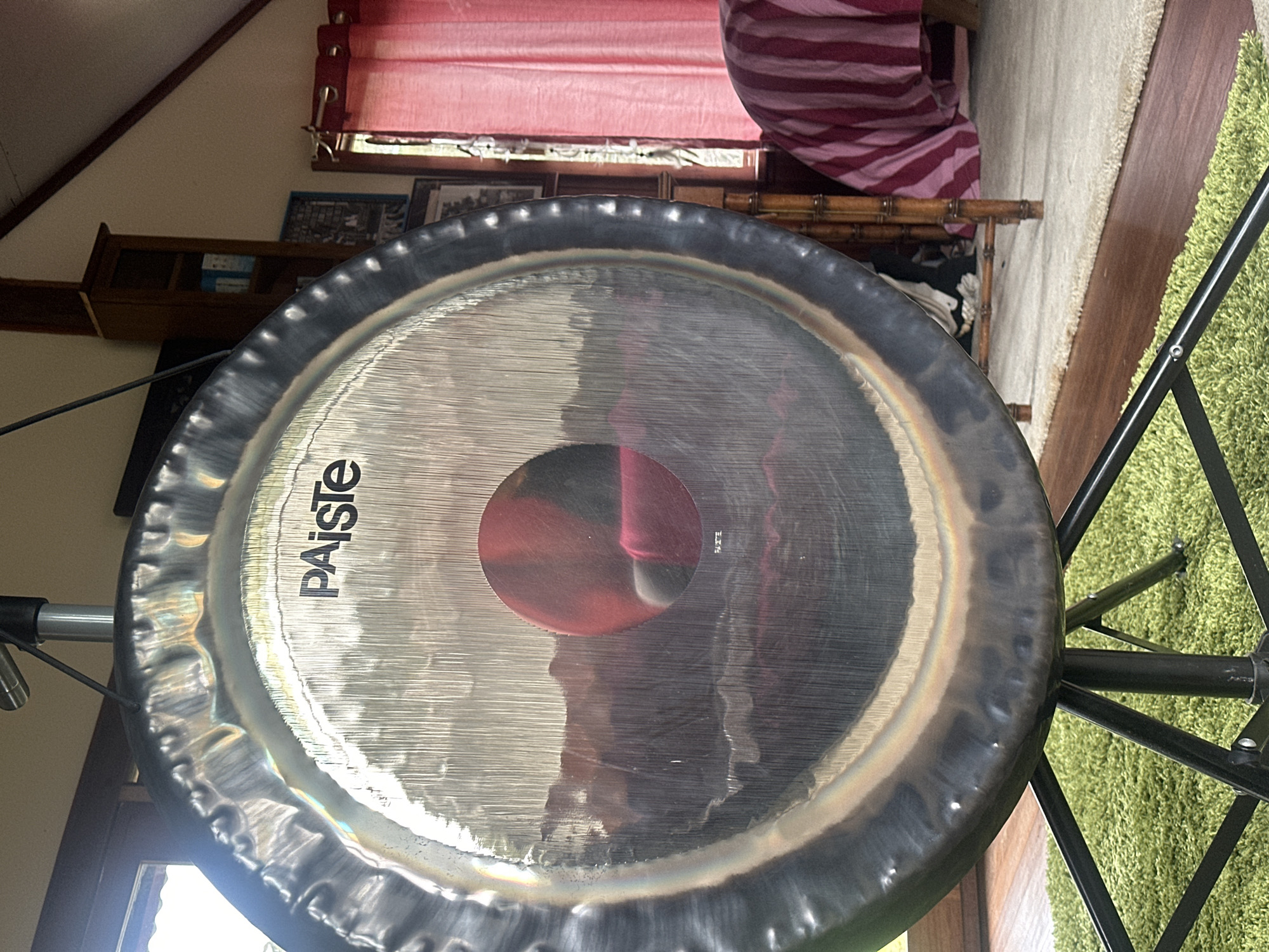 A large, circular Paiste gong is suspended from a stand, its metallic surface reflecting the surrounding environment. The gong is positioned in what appears to be a room with sloped ceilings and wooden floors. A green, shag rug is visible beneath the stand. In the background, red curtains are drawn across a window, and a wooden shelf unit with various items on it is present. A piece of furniture draped with a pink and white striped fabric is also visible. The scene is indoors, suggesting a personal space or studio. There is no indication of the city of Thora, Australia.