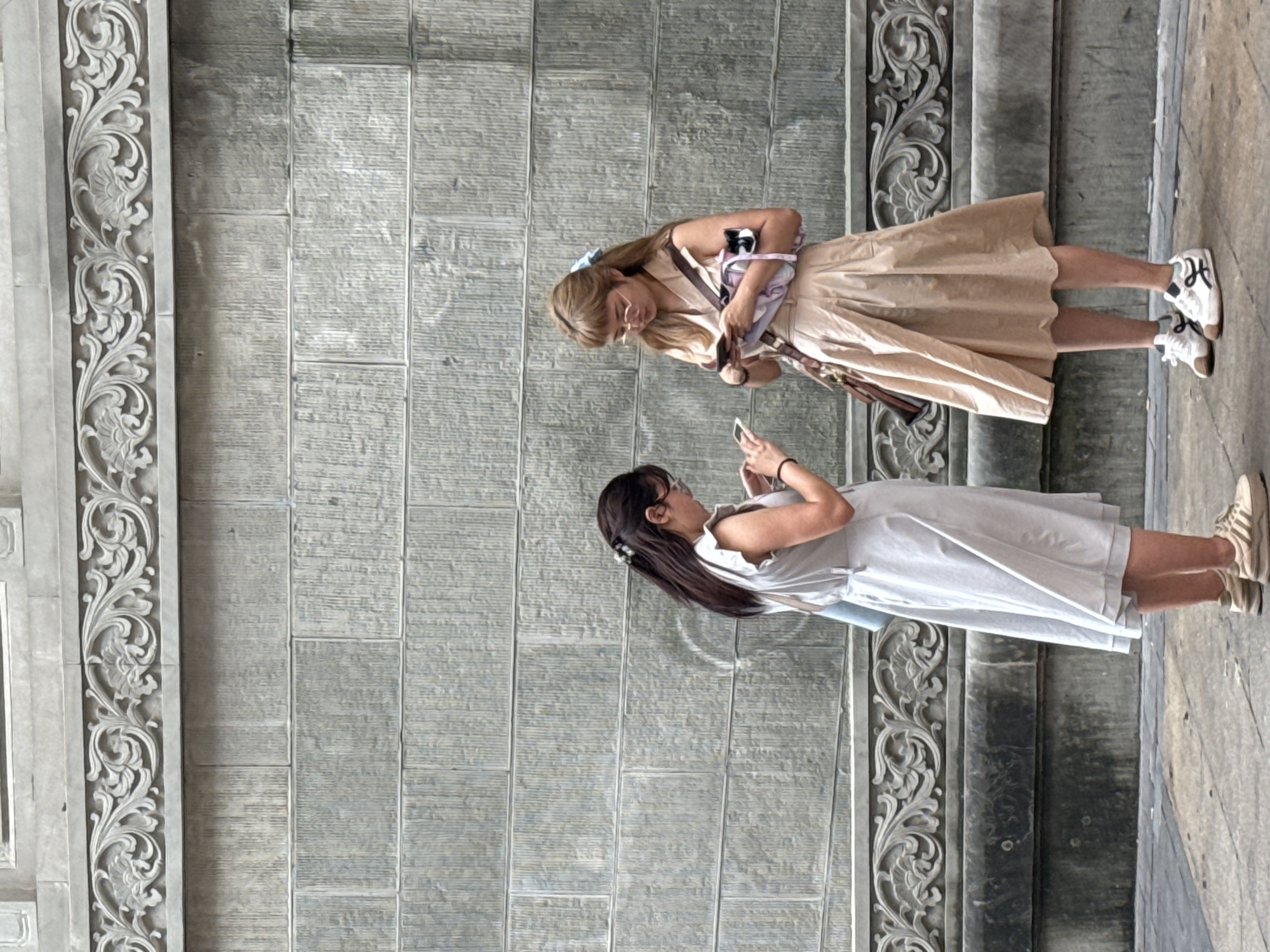 Two young women are standing in front of a textured gray stone wall in the Yuzhong District of China. The wall has intricate carved decorative borders at the top and bottom. Both women are looking down at their mobile phones. The woman on the left has long dark hair and is wearing a light-colored dress and sneakers. She has a light blue bag slung over her shoulder. The woman on the right has blonde hair with bangs and is wearing a tan dress and white sneakers. She is carrying a brown crossbody bag and a pink and white item in her left arm, which appears to be a small bag or pouch with a stuffed toy. They are both engaged with their phones, seemingly in a casual setting.