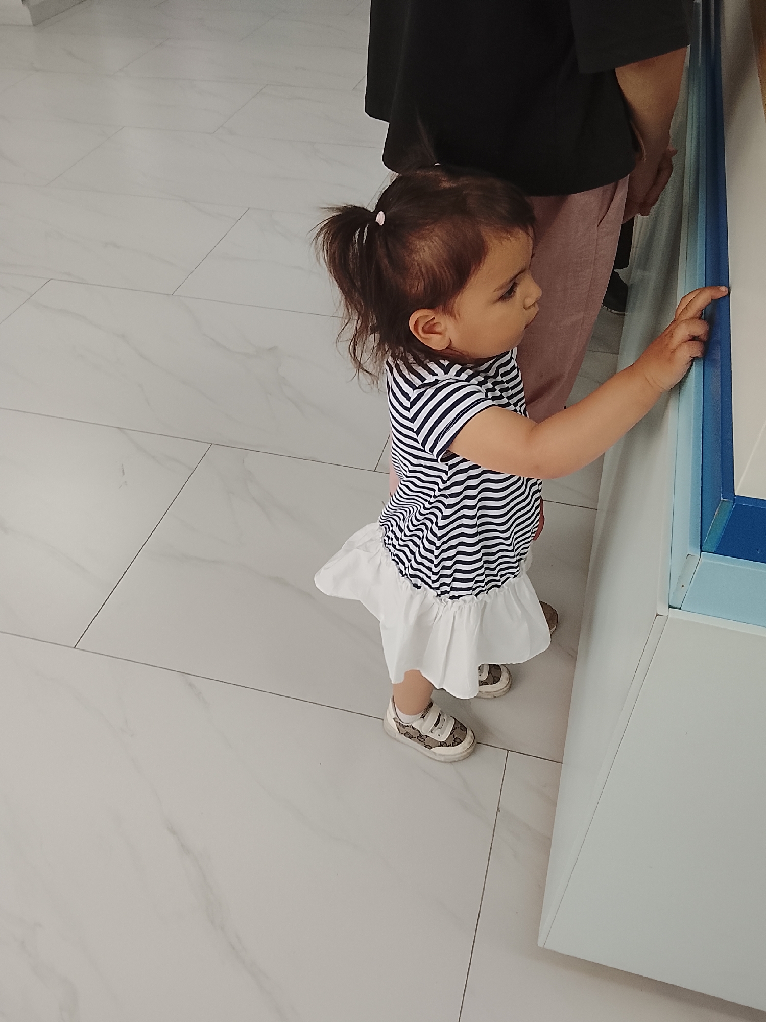 A young child with dark hair tied in a ponytail, wearing a navy and white striped shirt with a ruffled white skirt and patterned sneakers, stands facing a display case. The child's right hand is extended, touching the blue edge of the display. Behind the child, partially visible, is an adult wearing a black t-shirt and pink pants. The scene takes place indoors, on a polished white tiled floor with a marble pattern.
