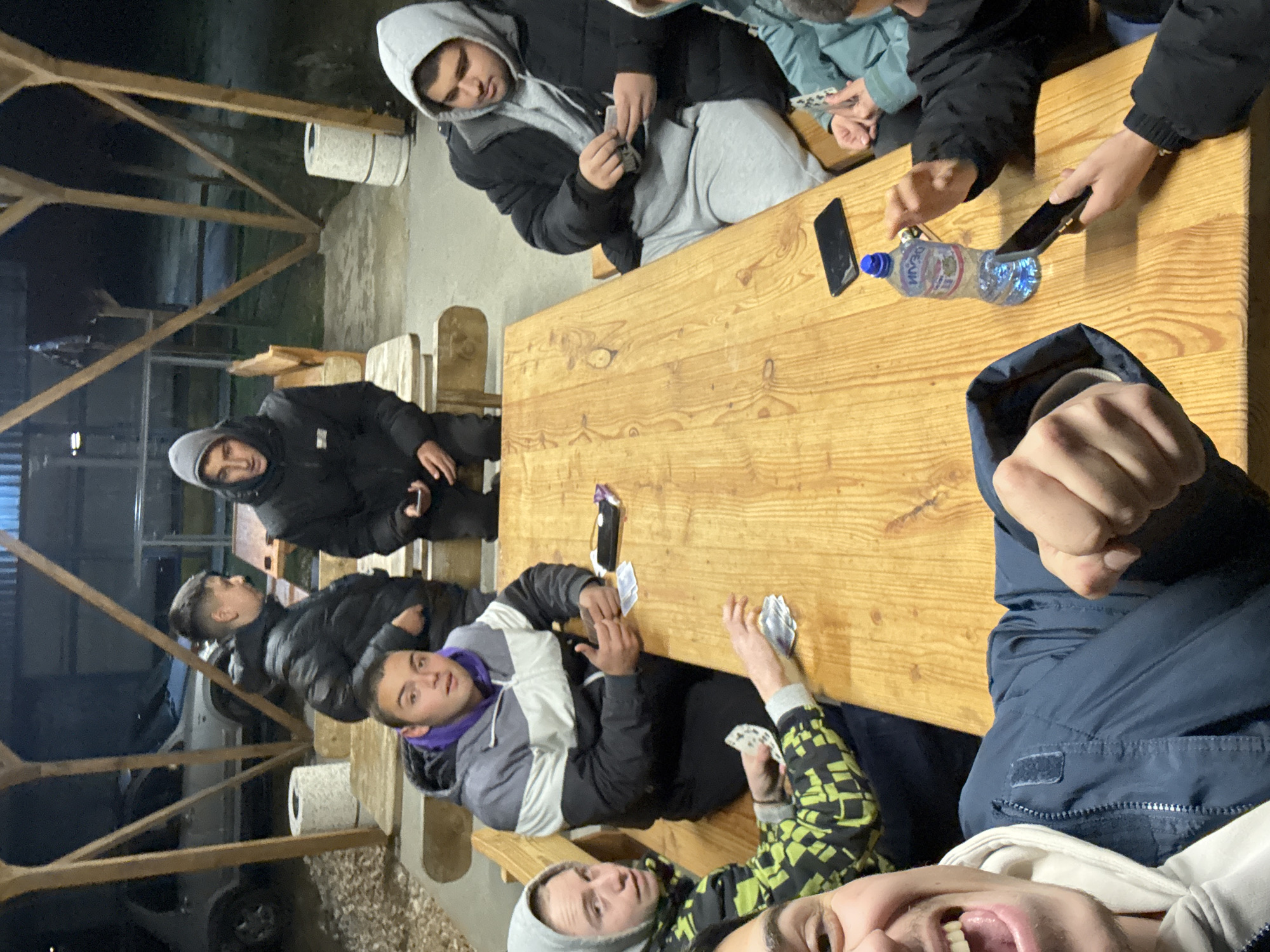 A medium shot shows a group of young men around a wooden picnic table in what appears to be an outdoor shelter. The scene is taken at dusk or night.
Seven young men are visible in the frame, most wearing hoodies and jackets. Some are holding or playing with cards, while others are looking at their phones. A water bottle and two phones are on the table.
The picnic table is made of light-colored wood. The outdoor shelter has wooden beams and posts, with a glimpse of a car visible in the background. A roll of what might be paper towels or toilet paper is on a nearby support post.
The location and city are unidentified.
